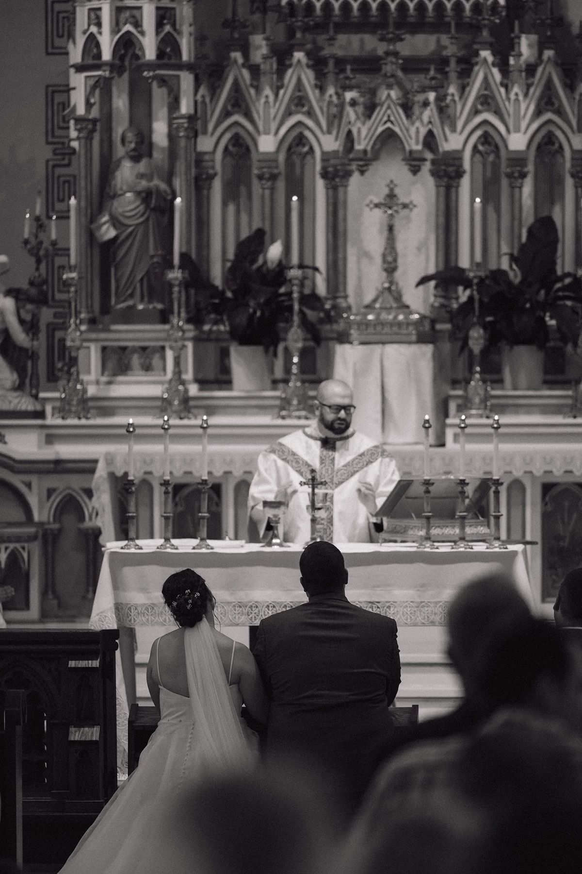 Black and white photo of a bride and groom kneeling before a priest at a Catholic church wedding ceremony altar.