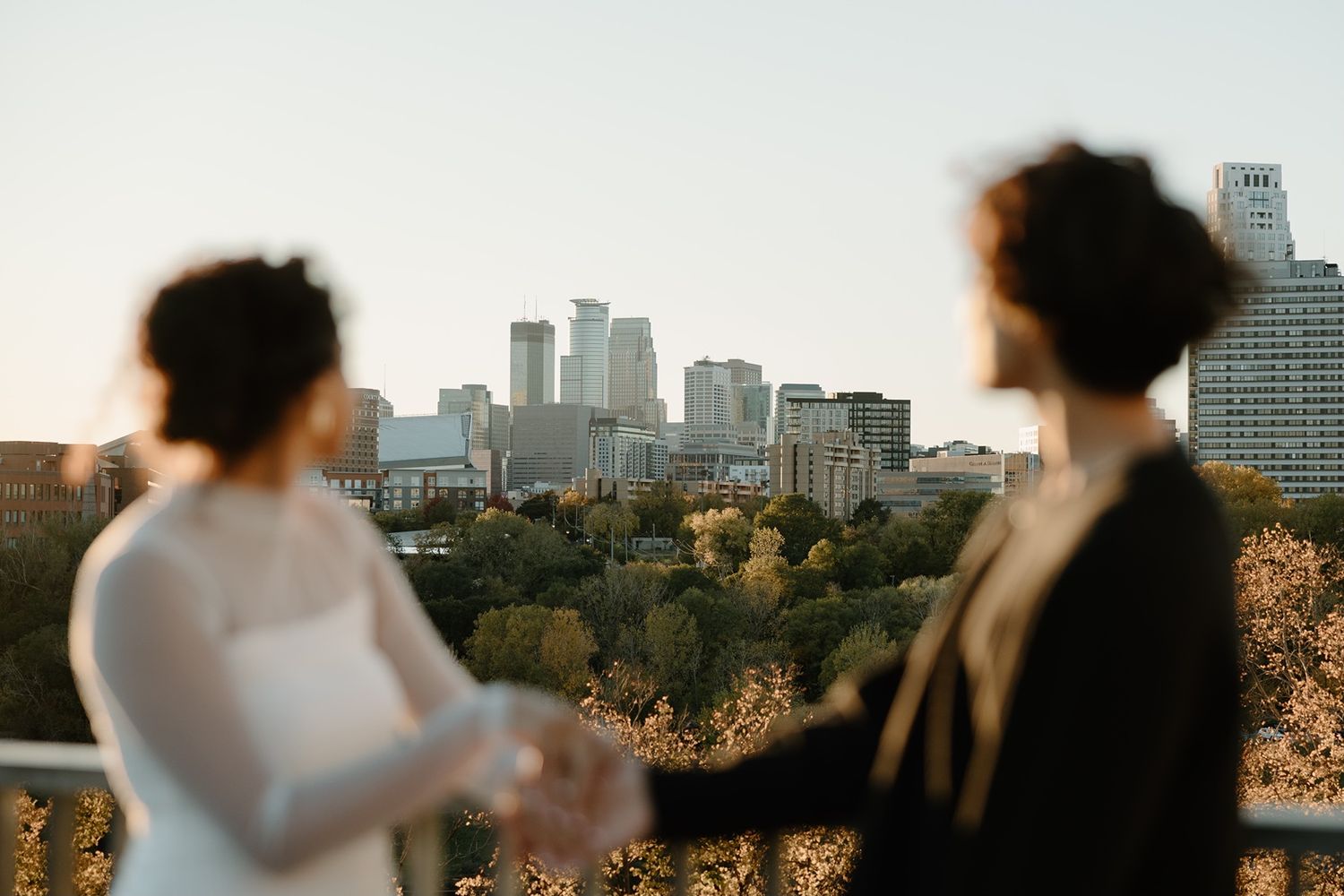 A couple holds hands on a rooftop with a city skyline and autumn trees in the background.