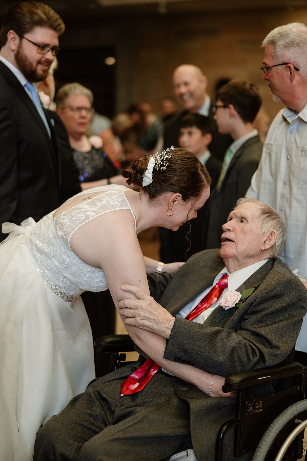 A tender moment between a bride in white and elderly person in wheelchair during reception.