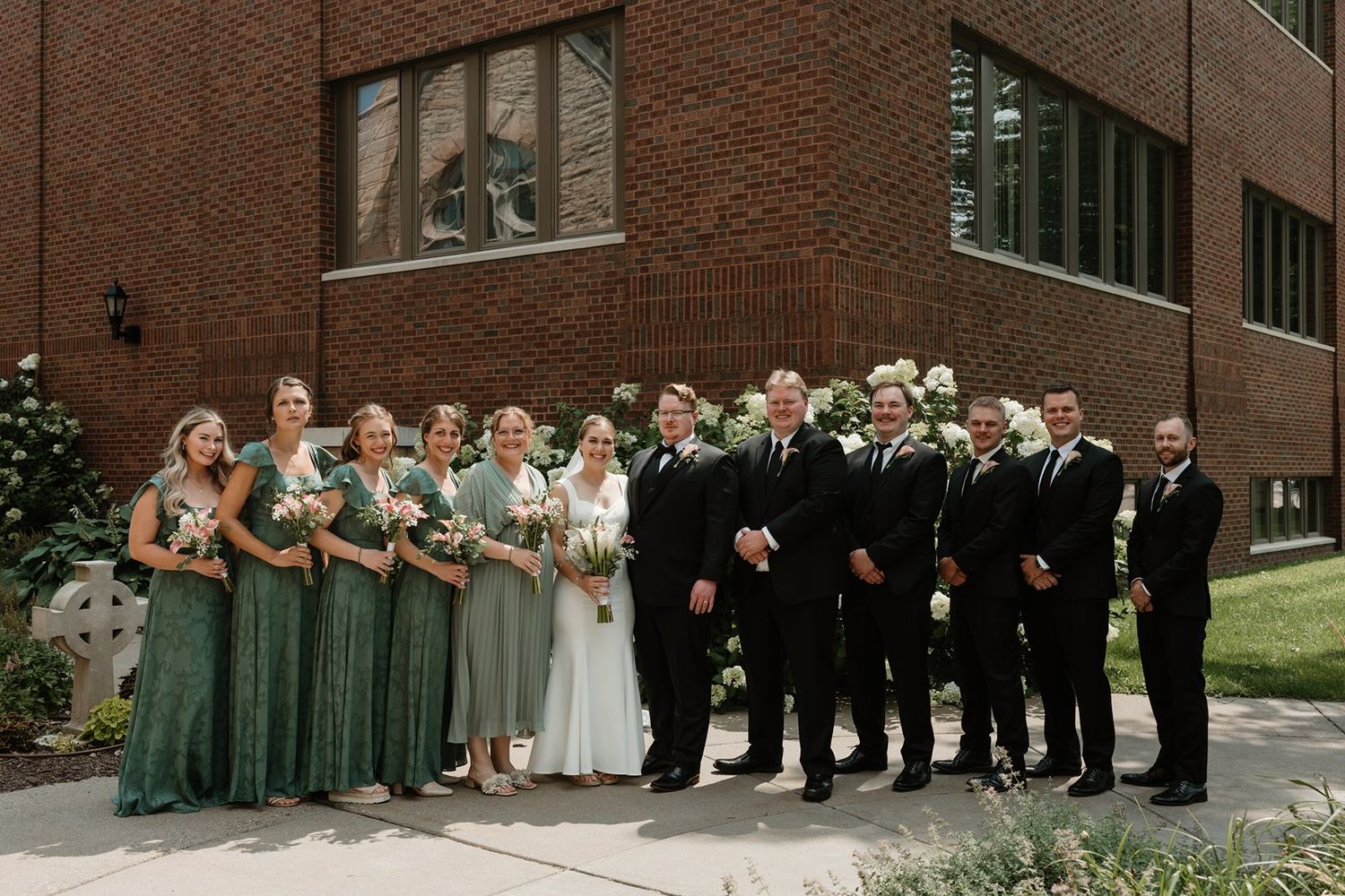 Wedding party in formal attire poses outdoors against a brick building backdrop.
