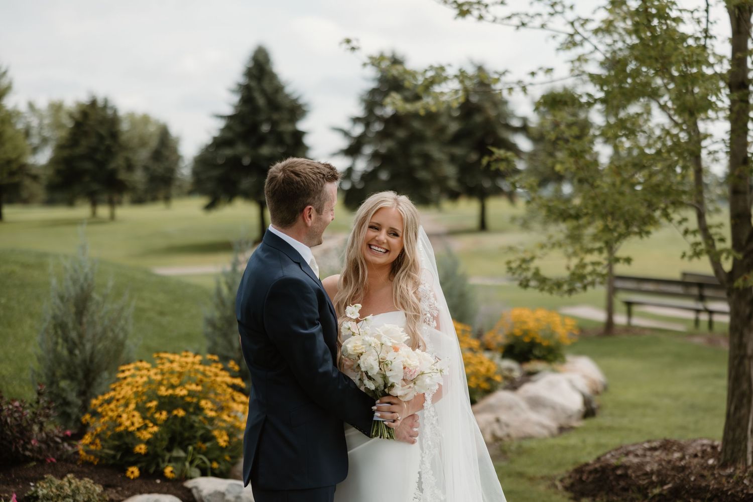 Newlywed couple stands together in a garden setting with yellow flowers and evergreen trees in the background.
