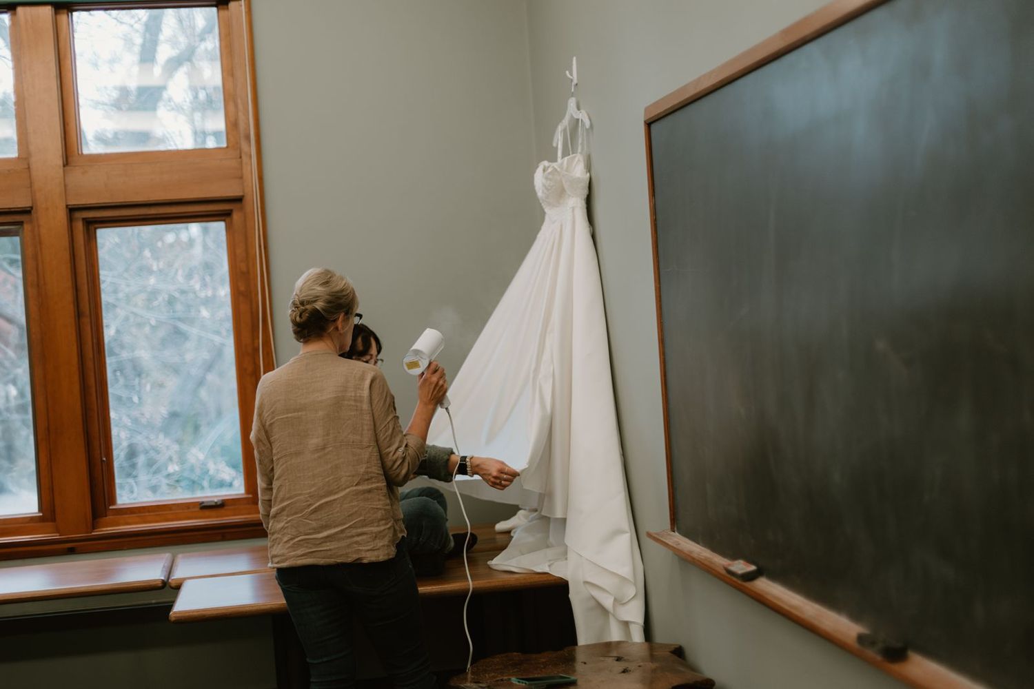 Wedding dress being prepared in classroom setting with chalkboard and window.