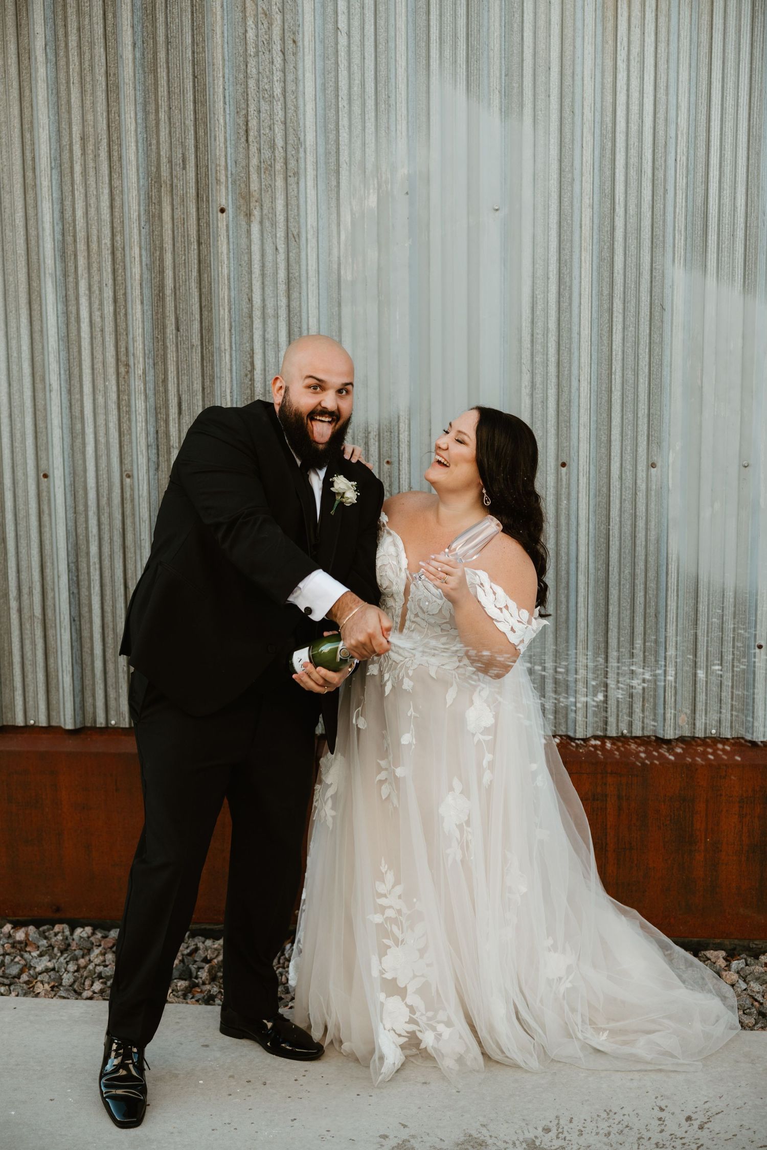 A joyous couple laughs together against a corrugated wall on their wedding day, wearing formal black suit and white gown.