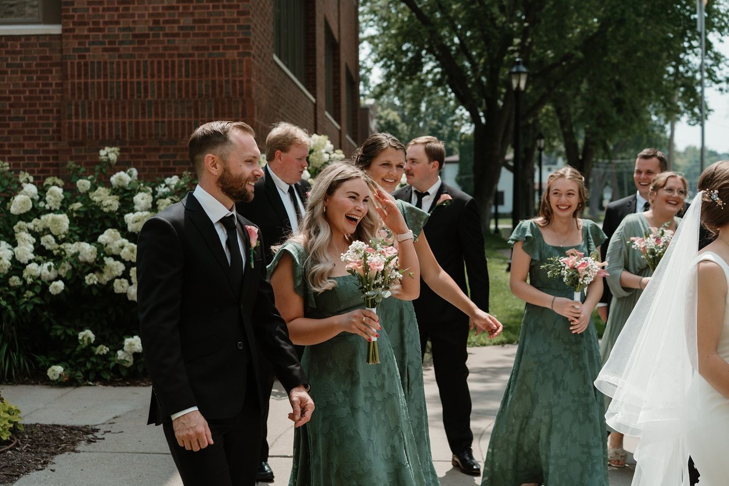 Wedding party celebrates with champagne outside brick building in matching sage green dresses.