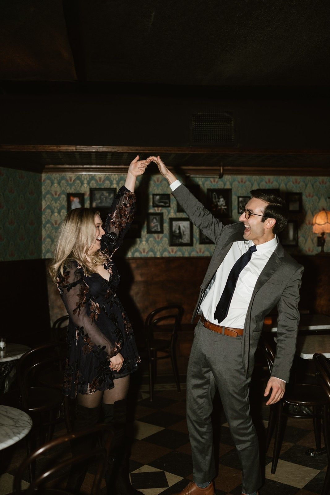 Two people dancing together in a dimly lit vintage-style speakeasy with patterned wallpaper and moody lighting.