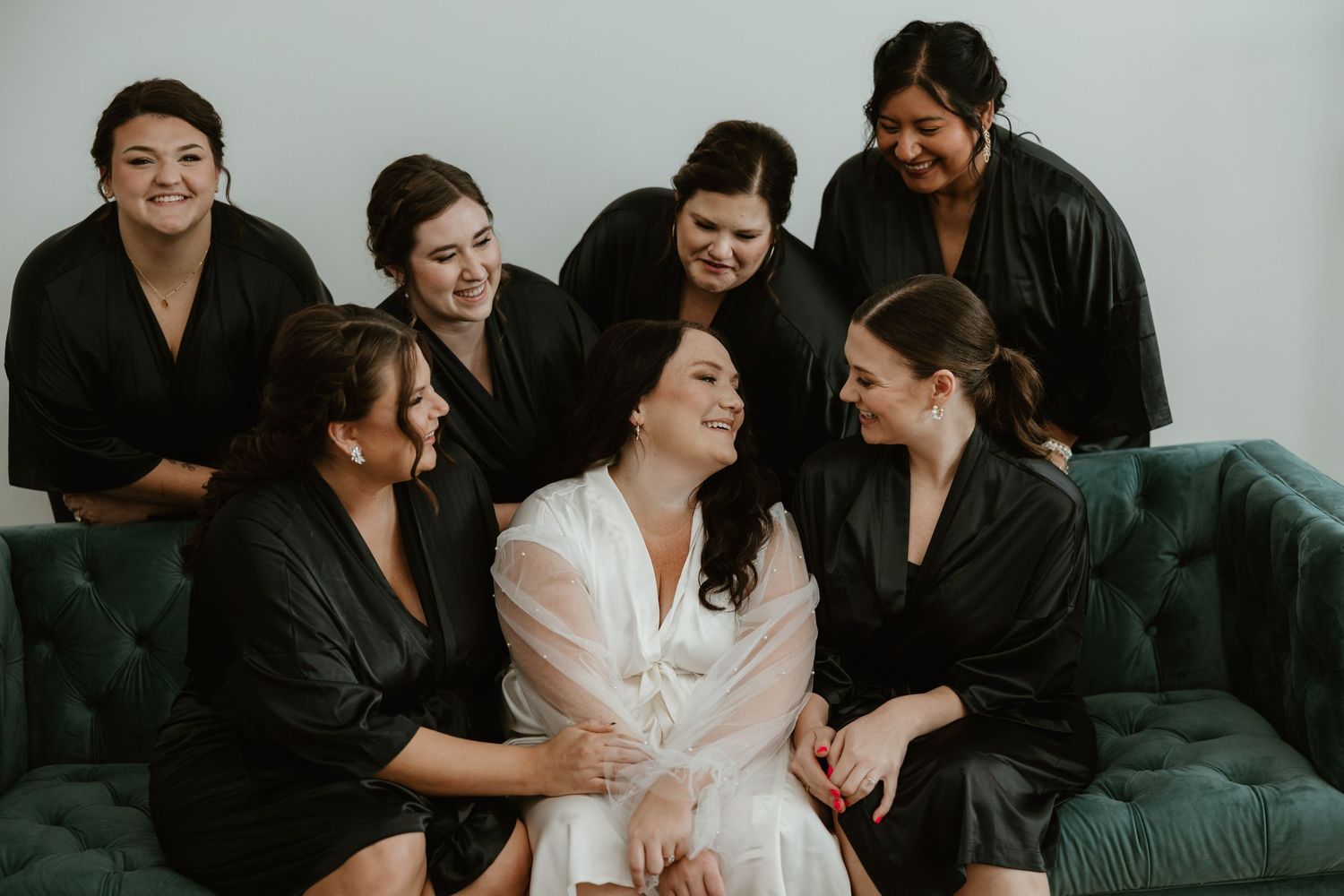 A group wearing black robes surrounds someone in white silk robe sitting on a teal velvet couch while sharing laughs together.