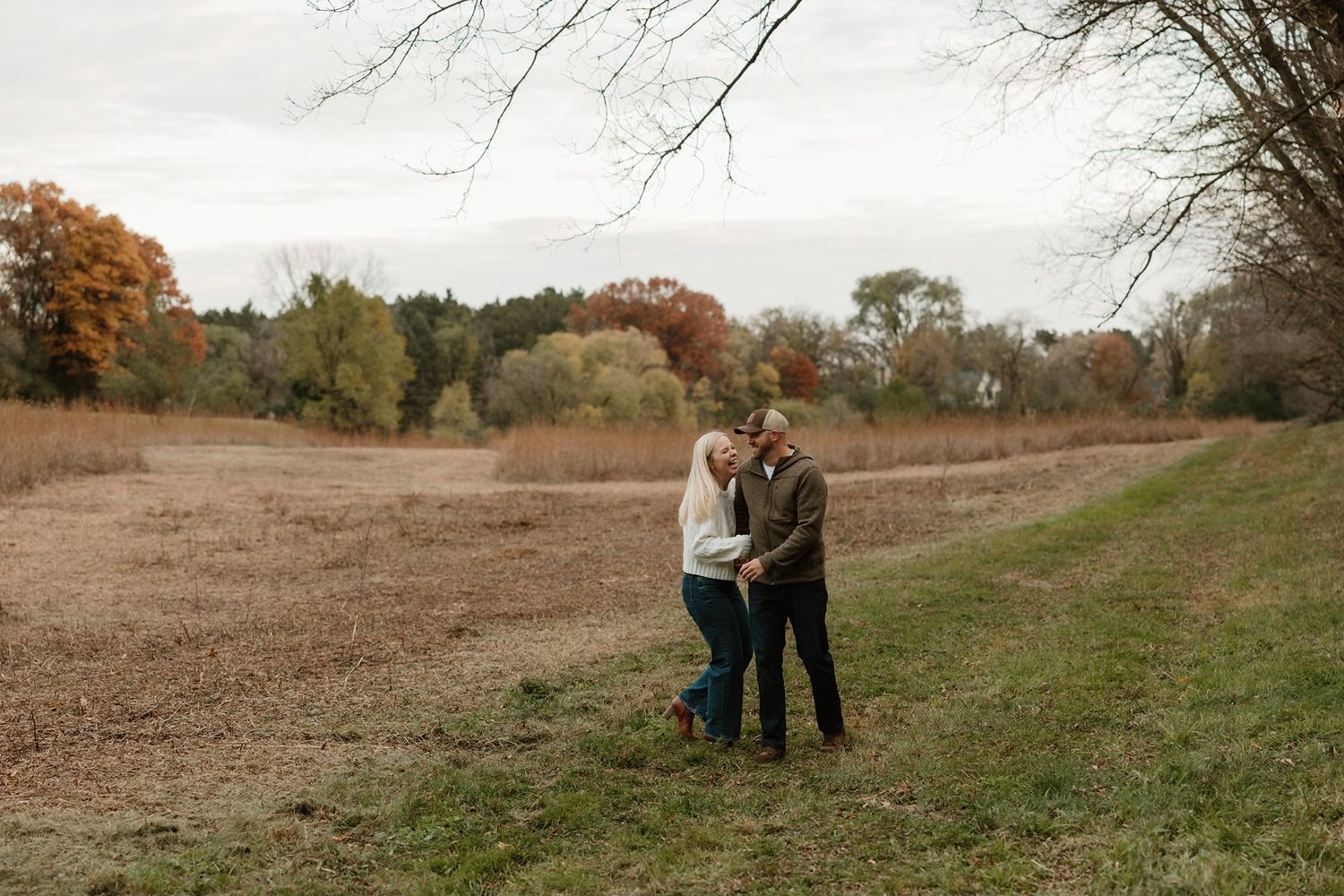 Couple embracing in an autumn field surrounded by colorful fall foliage and bare trees on a cloudy day.