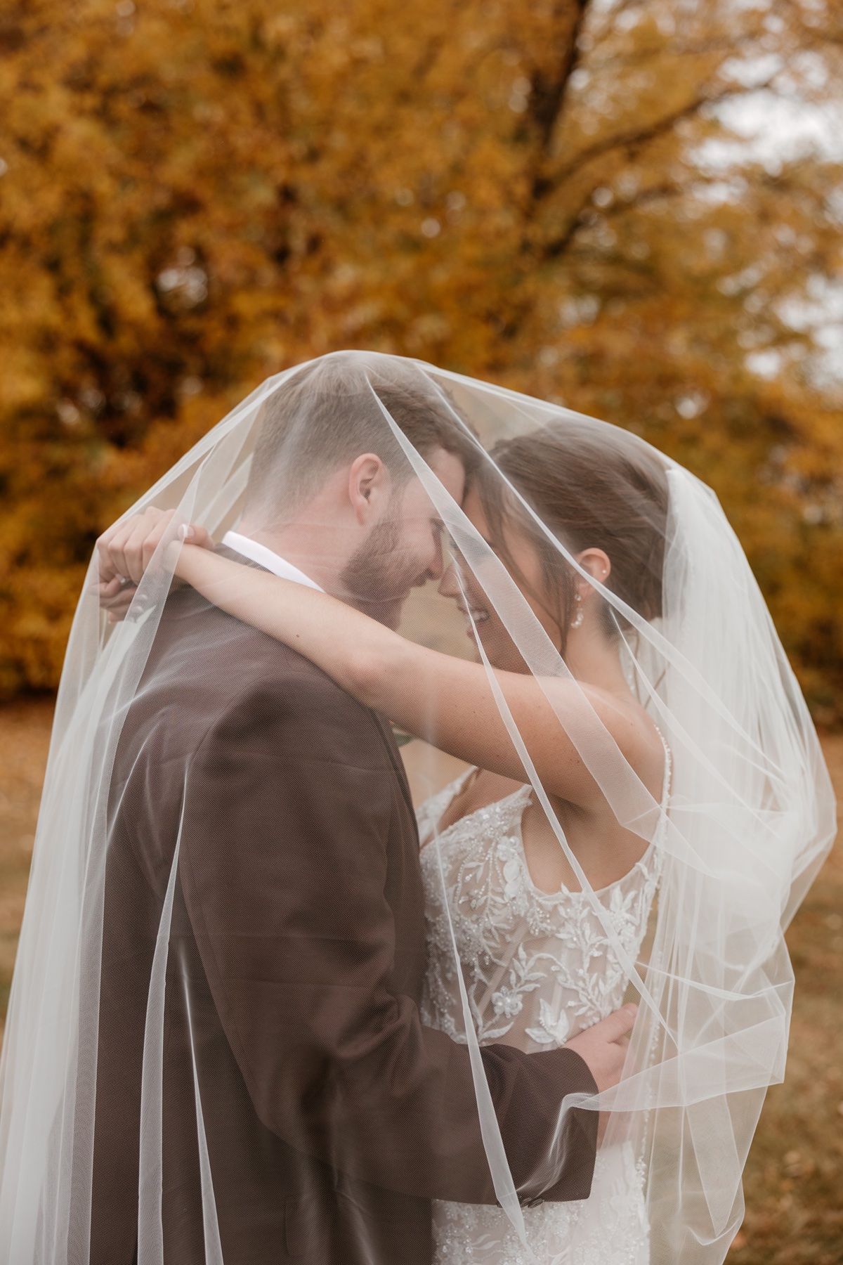 Bride and groom sharing a romantic moment under a white veil with golden autumn trees in the background.