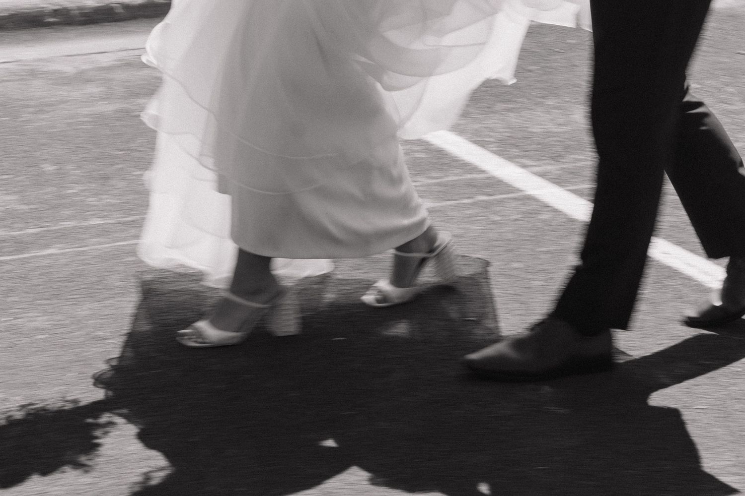 Black and white photo of bride and groom walking, showing wedding dress hem and formal shoes on pavement.