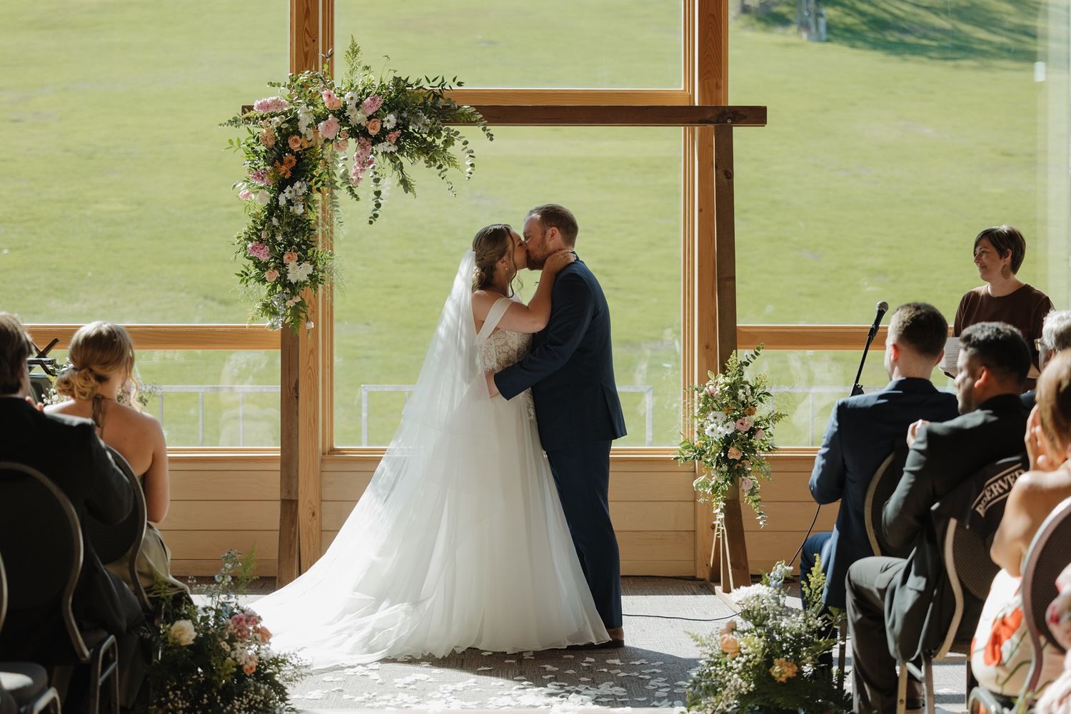 Bride and groom share first kiss at wedding altar with floral arch and scenic green field backdrop.