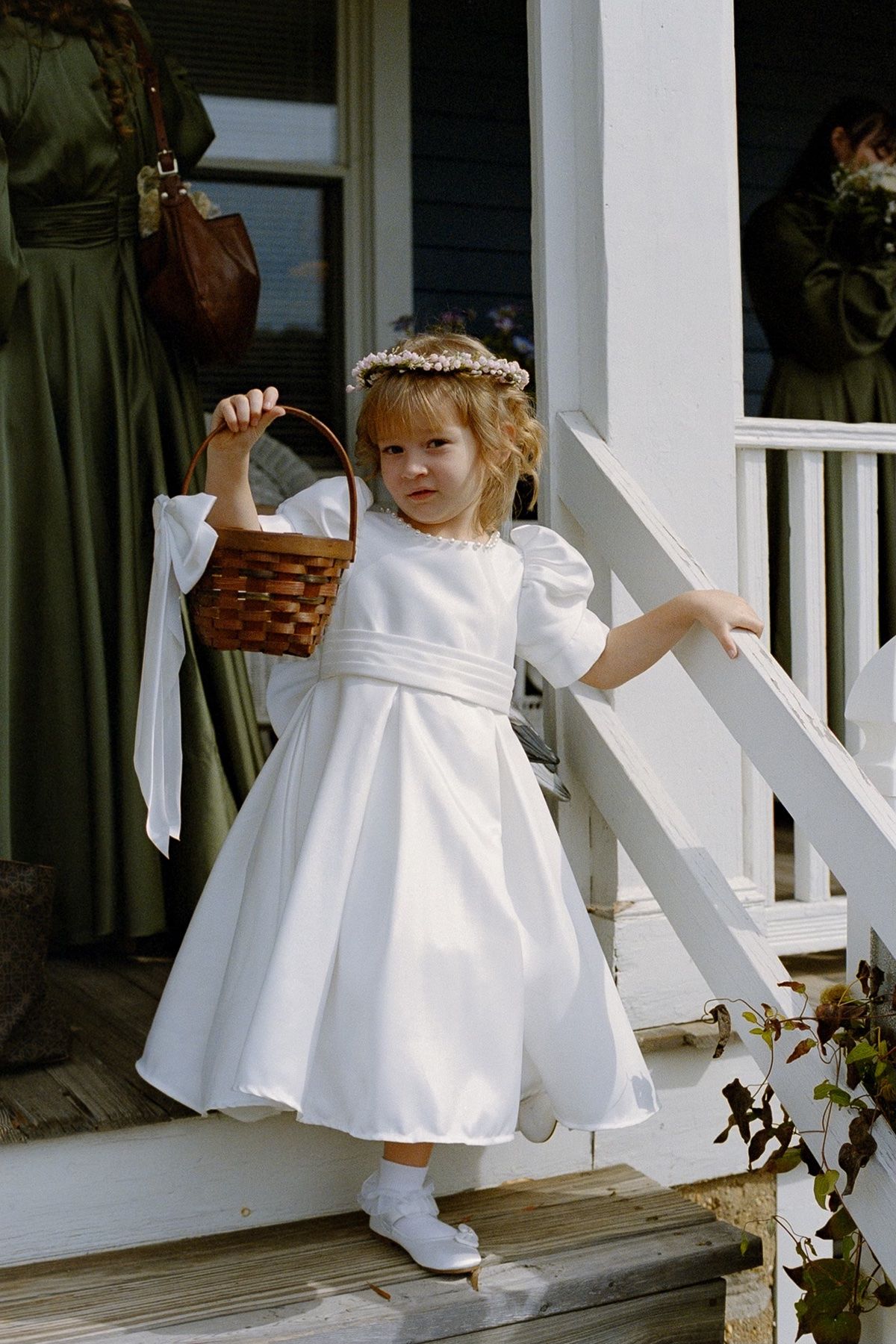 Young flower girl in white dress holding wicker basket on porch steps at wedding ceremony.