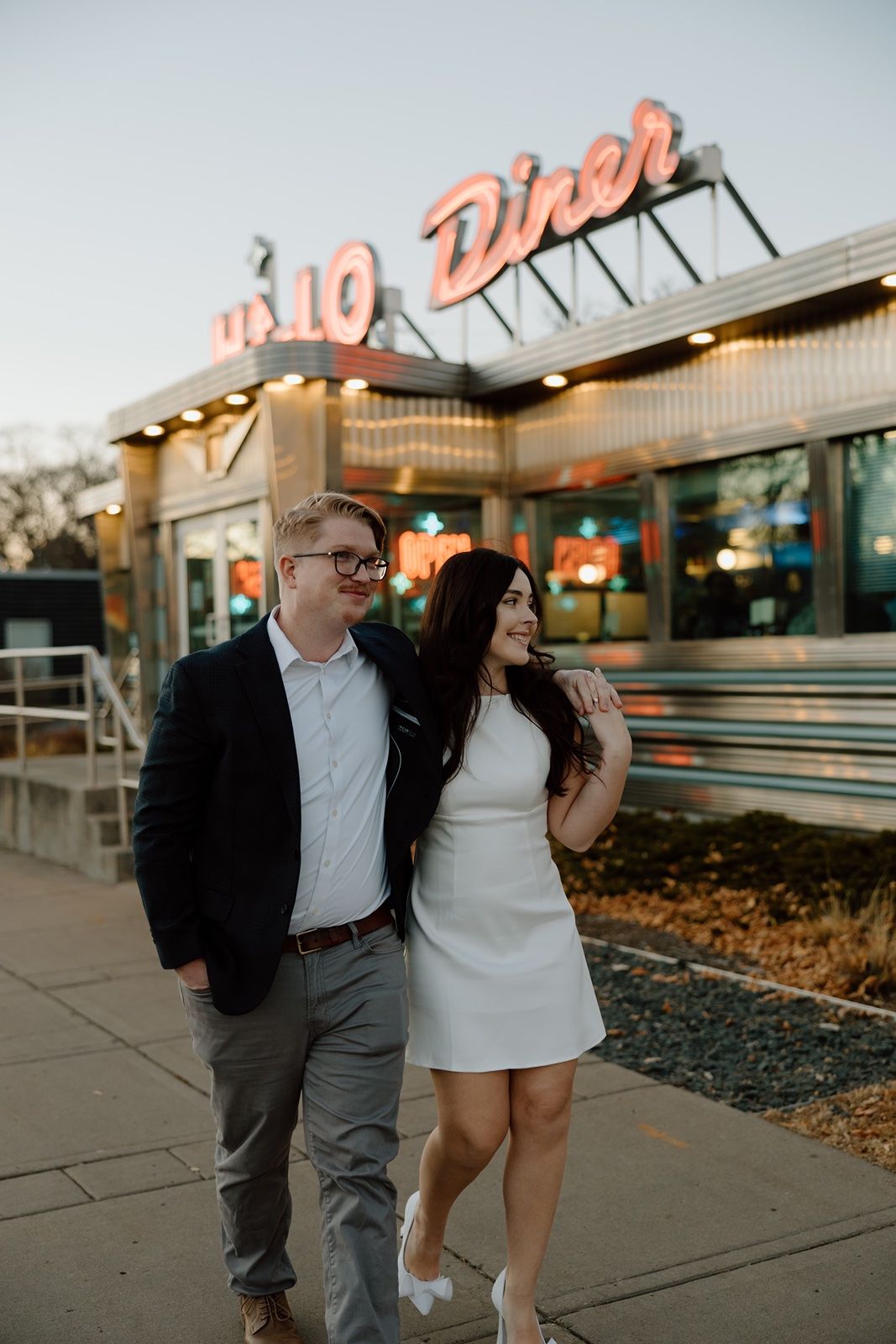 Couple poses outside a retro neon-lit Hi-Lo diner in Minneapolis at dusk, woman in white dress, man in dark blazer.