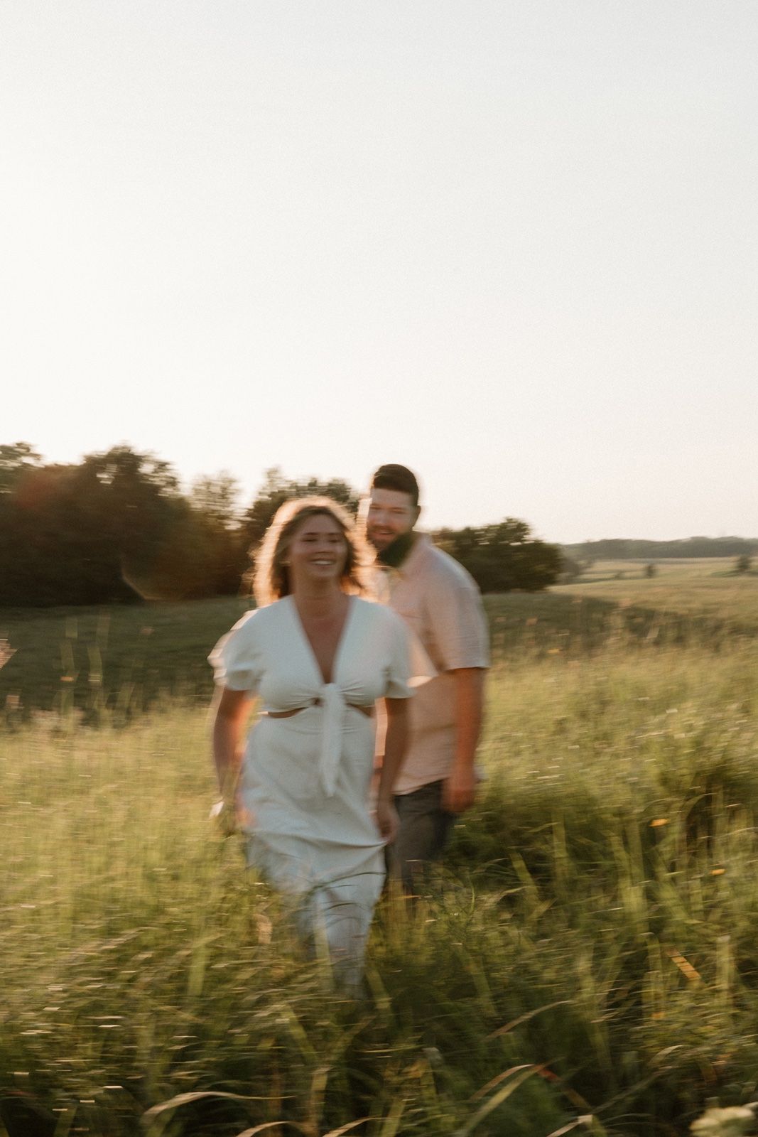 A couple walks through a sunlit green field at golden hour, woman in white dress smiling, bearded man following behind.