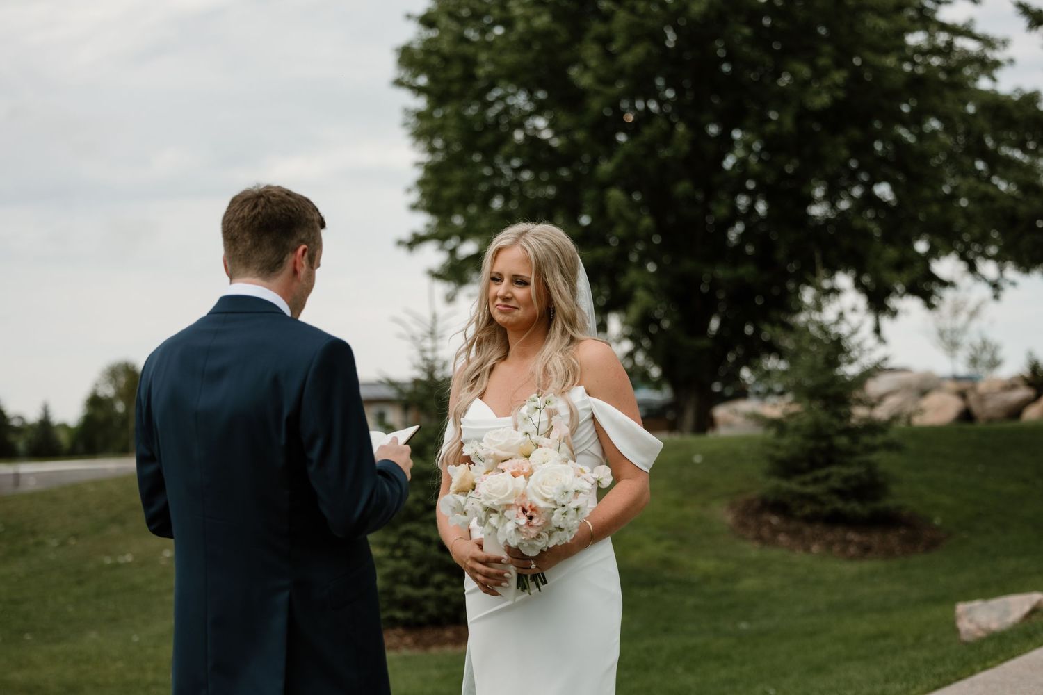 Couple in wedding attire embracing in a lush green outdoor setting at sunset.