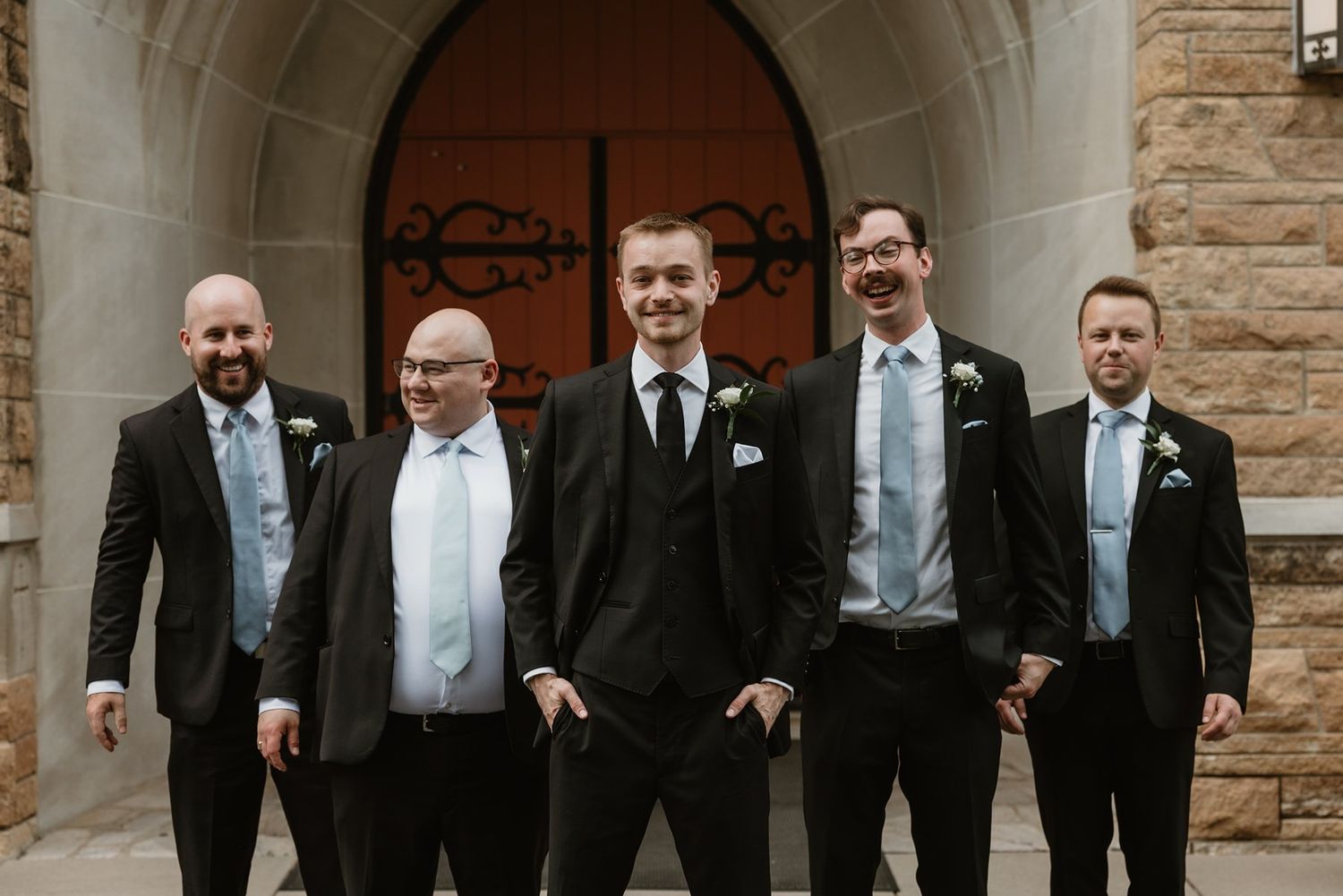 Five smiling groomsmen in black suits and white shirts pose together in front of a stone church archway.