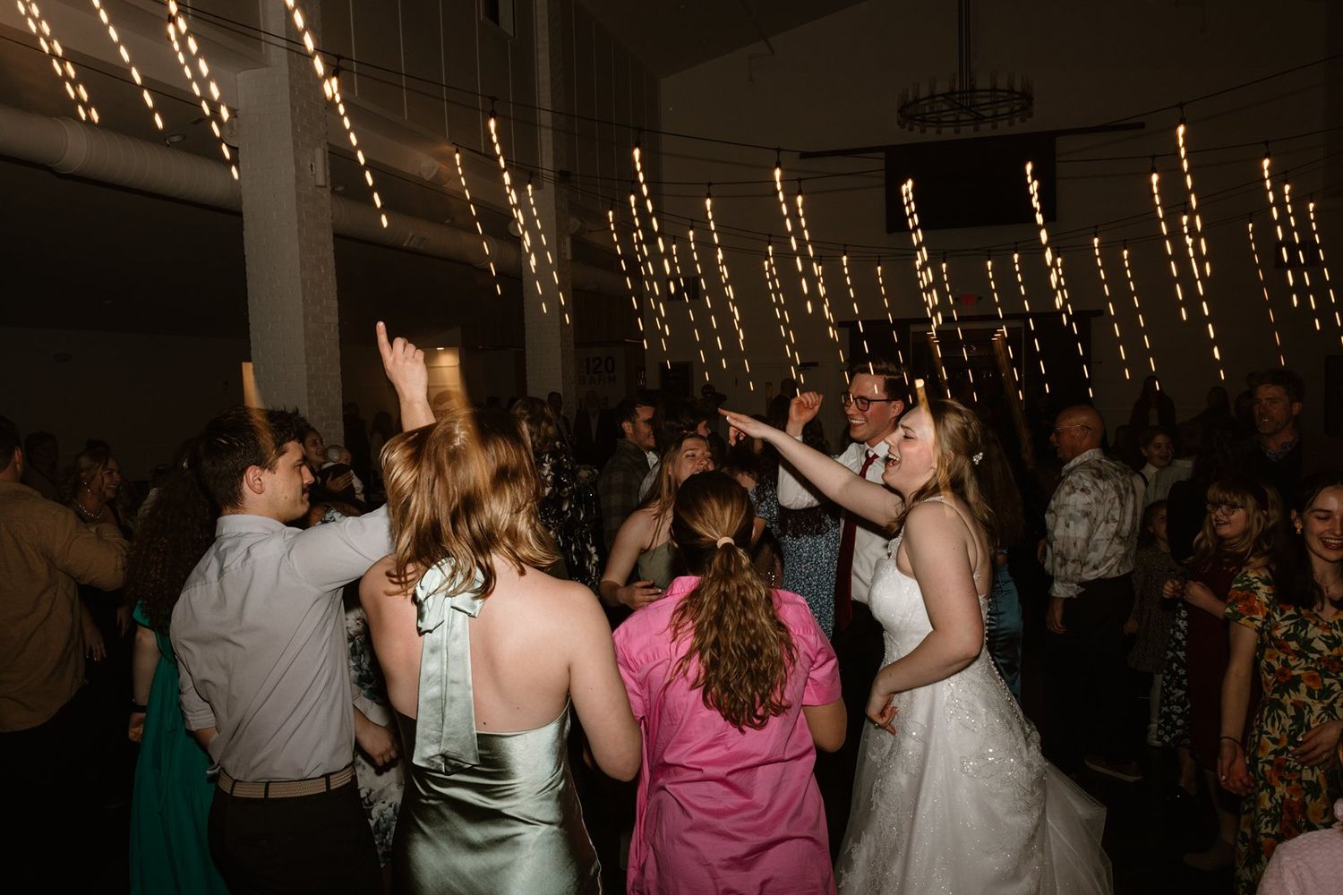 Wedding reception dance floor filled with guests celebrating under warm string light decorations.