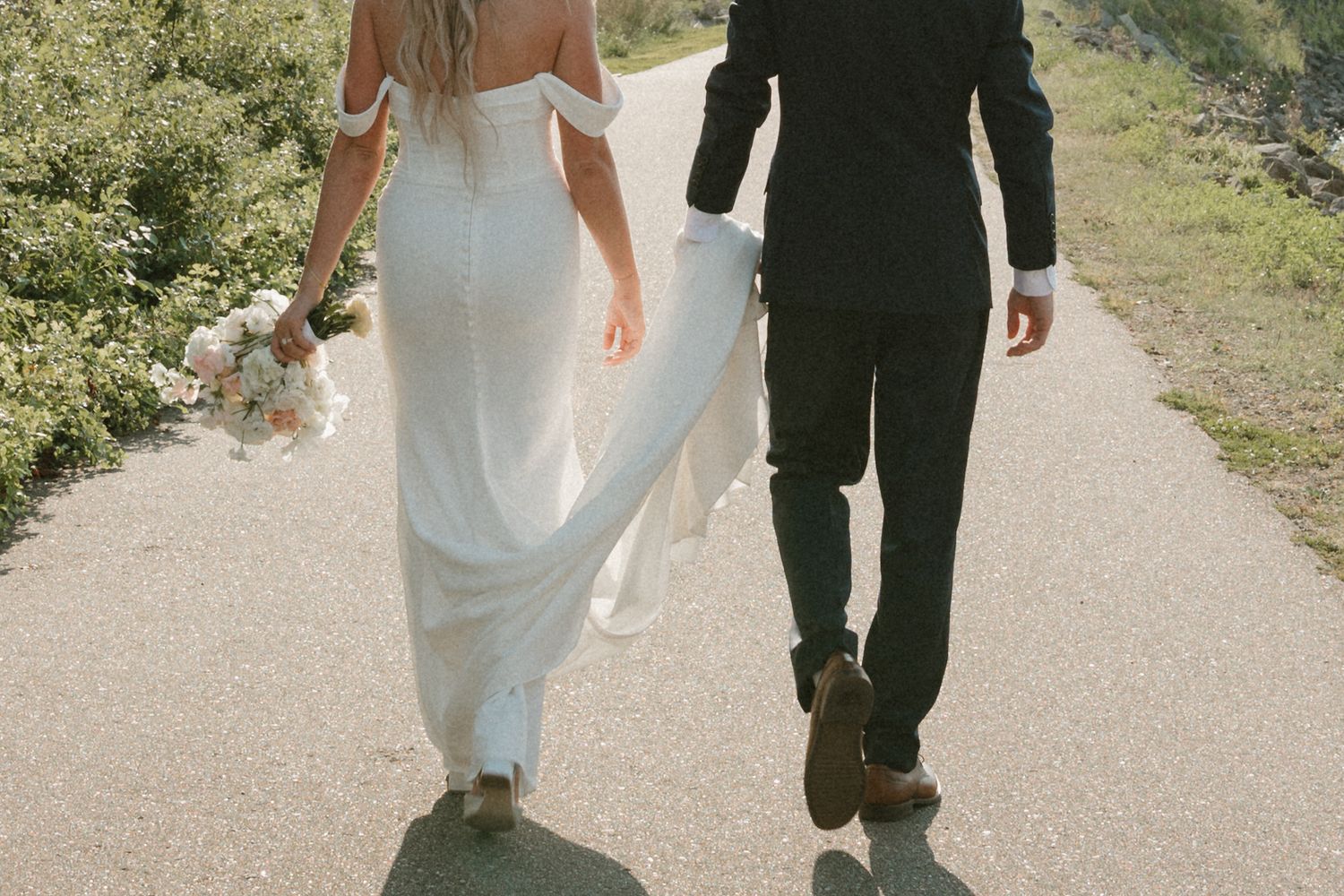A couple walks down a sunlit path on their wedding day, with the bride in white and groom in black.