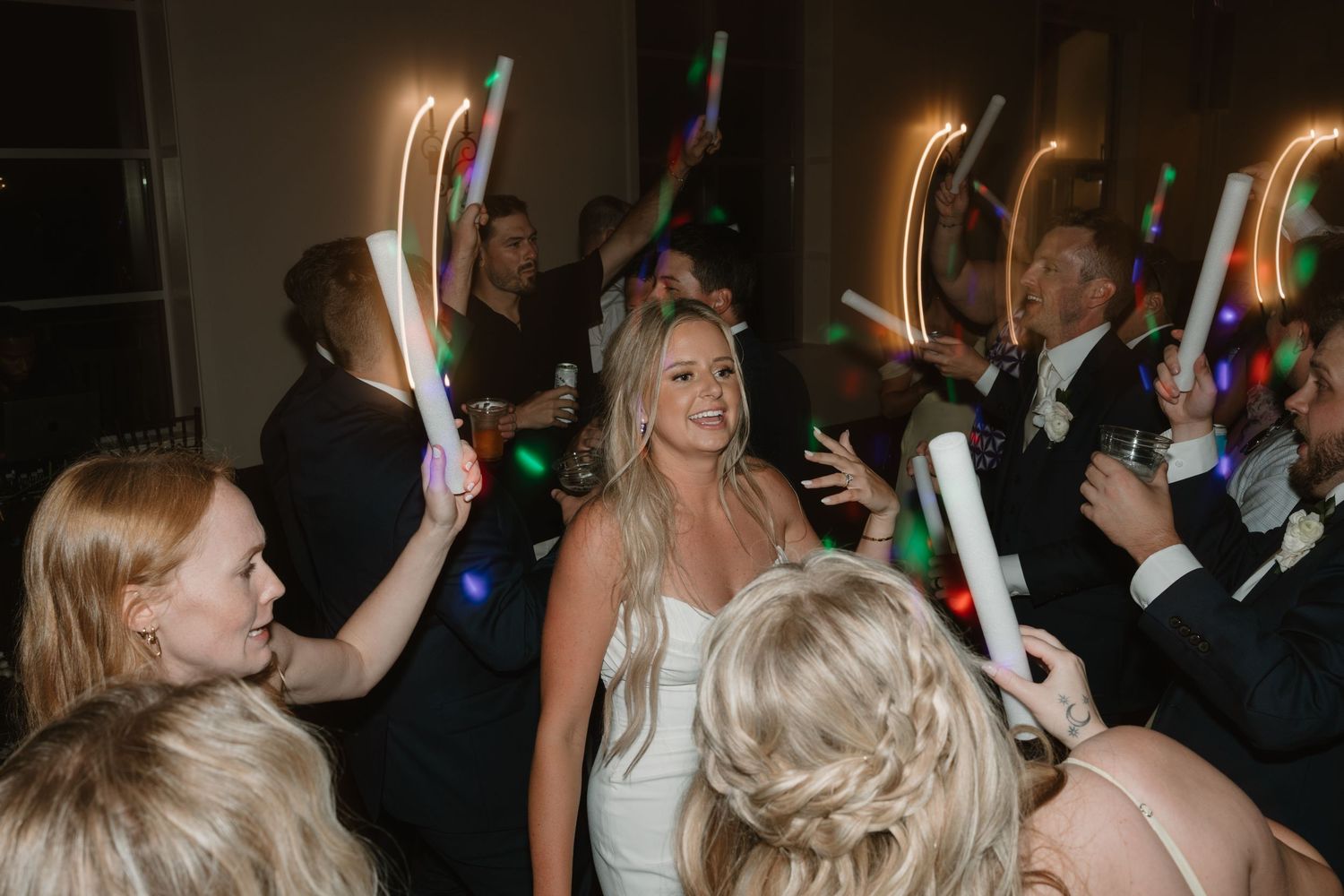 Wedding party dances with glowing neon light sticks creating colorful light trails in a dark reception venue.
