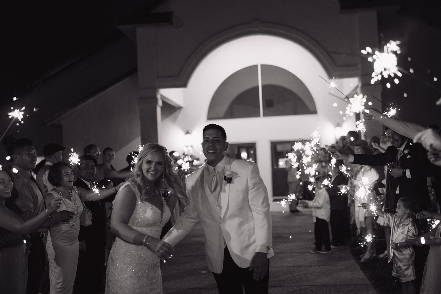 Couple exits their wedding venue through a crowd holding bright sparklers at night.