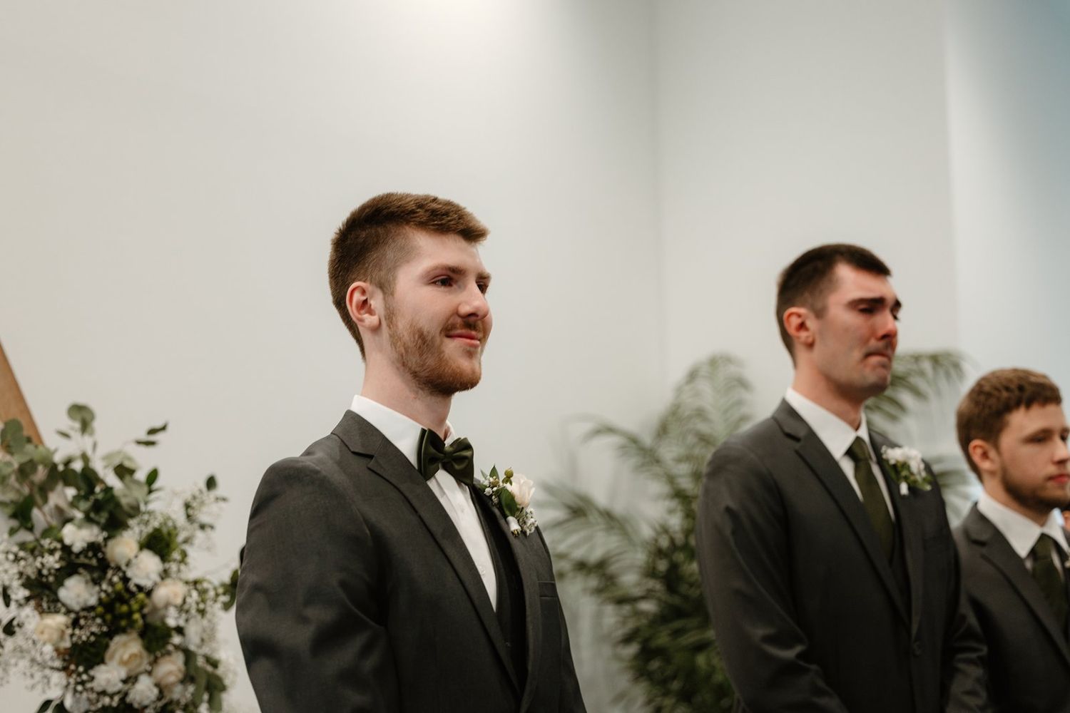 Wedding party members in dark suits stand together during an outdoor ceremony against a cloudy sky.