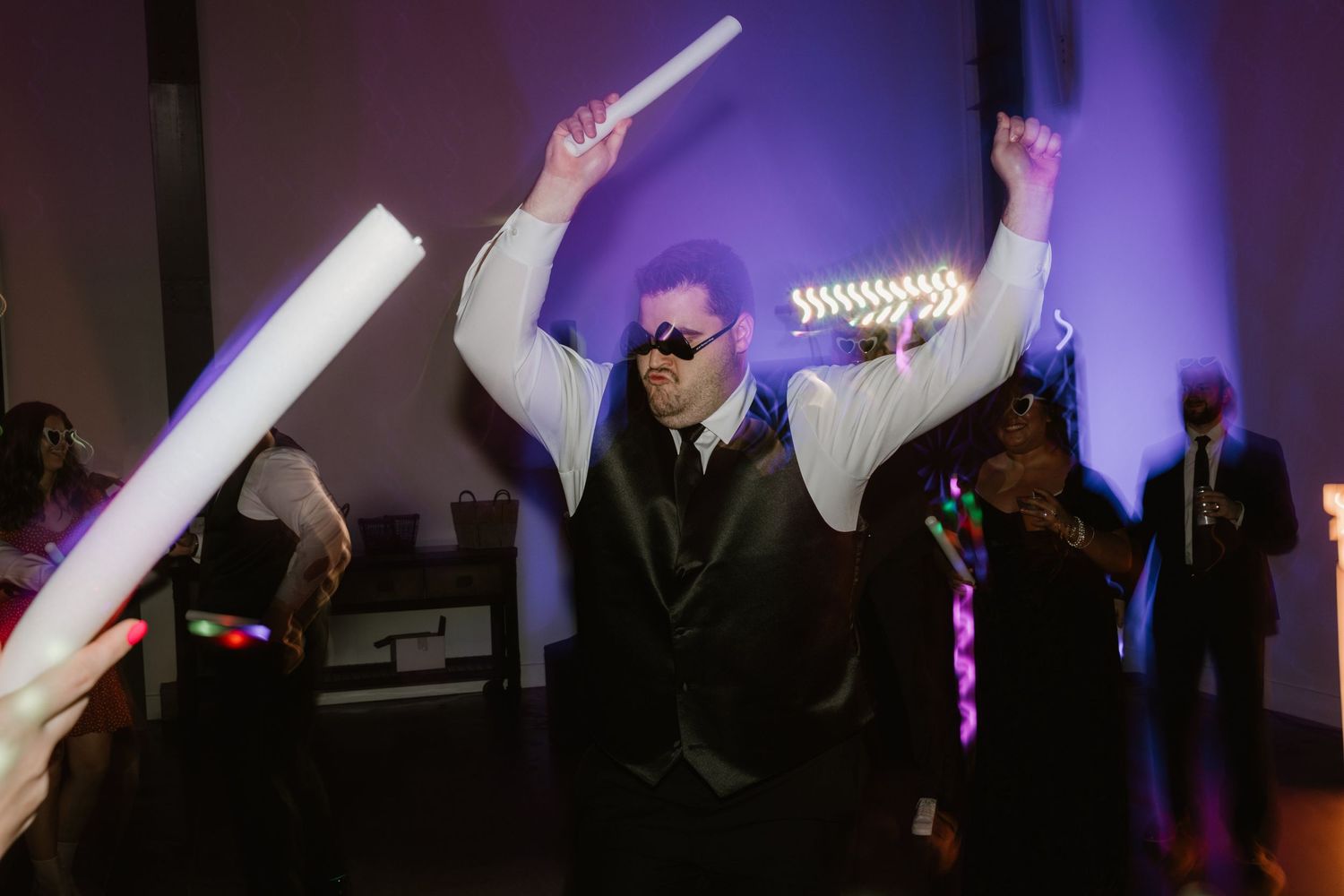 Partygoer in sunglasses and suit holding up glowing LED foam sticks while dancing at a nighttime celebration.