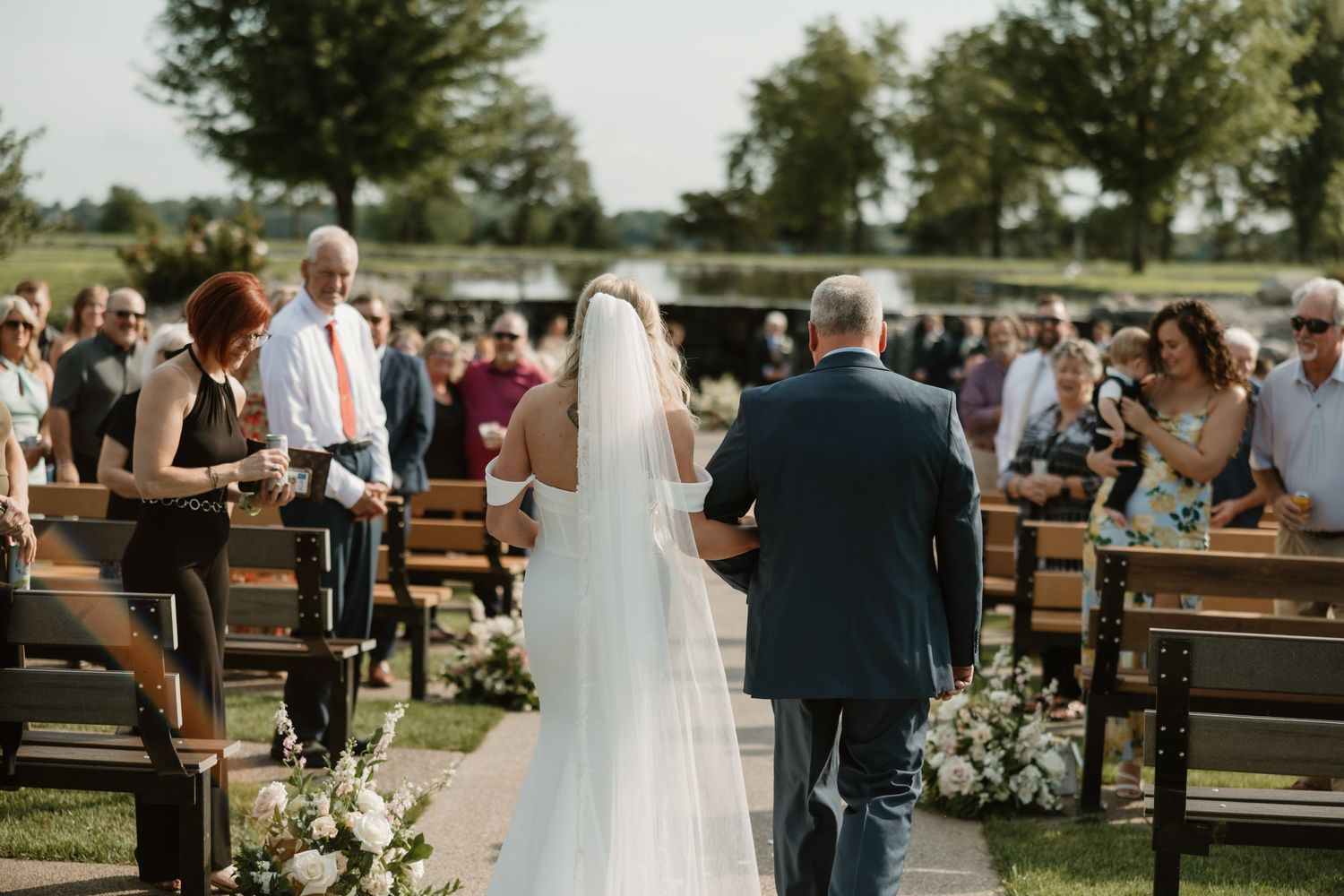 An outdoor wedding ceremony with wooden benches and floral arrangements along the aisle during a sunny afternoon.