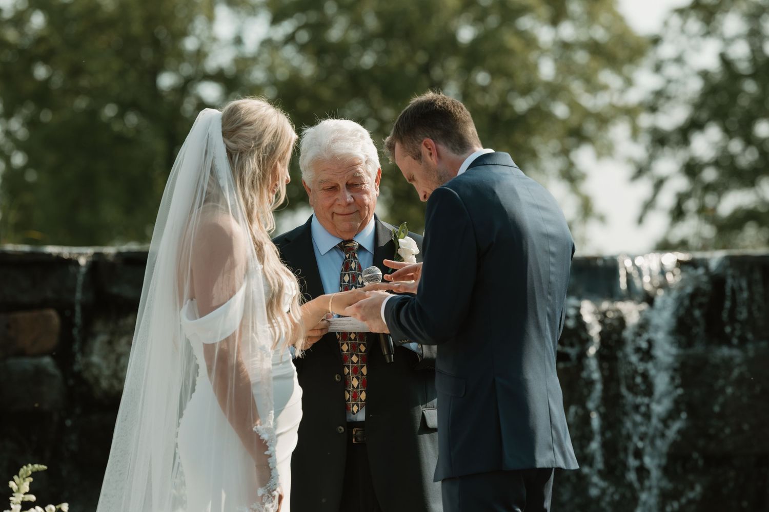 An outdoor wedding ceremony takes place in front of a stone wall and waterfall as an officiant leads the couple's vows.