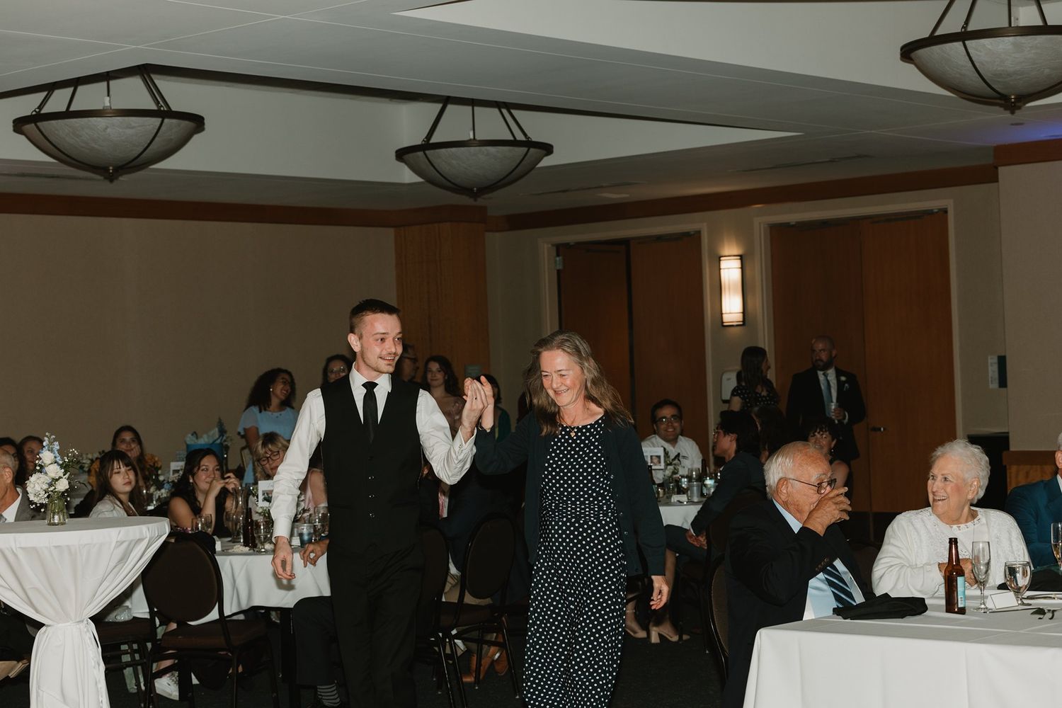 Wedding reception guests dance and celebrate in an elegant ballroom with overhead lighting fixtures.
