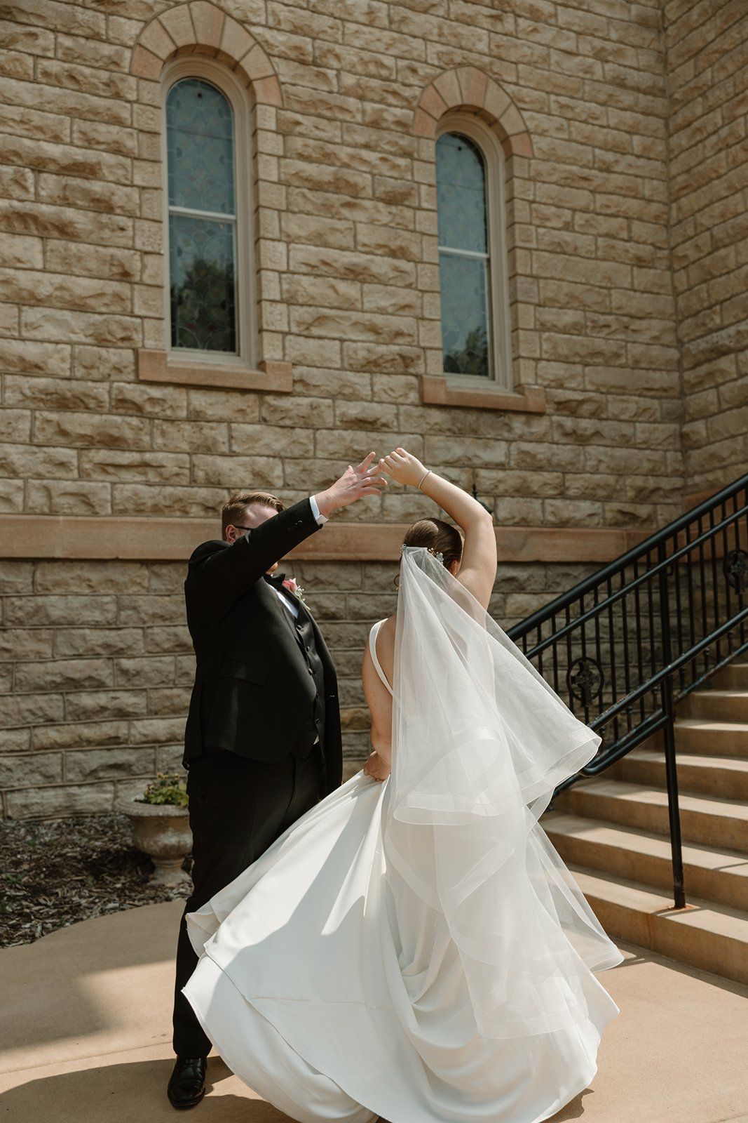 A couple dances elegantly on church steps with flowing white dress and veil against stone wall architecture.