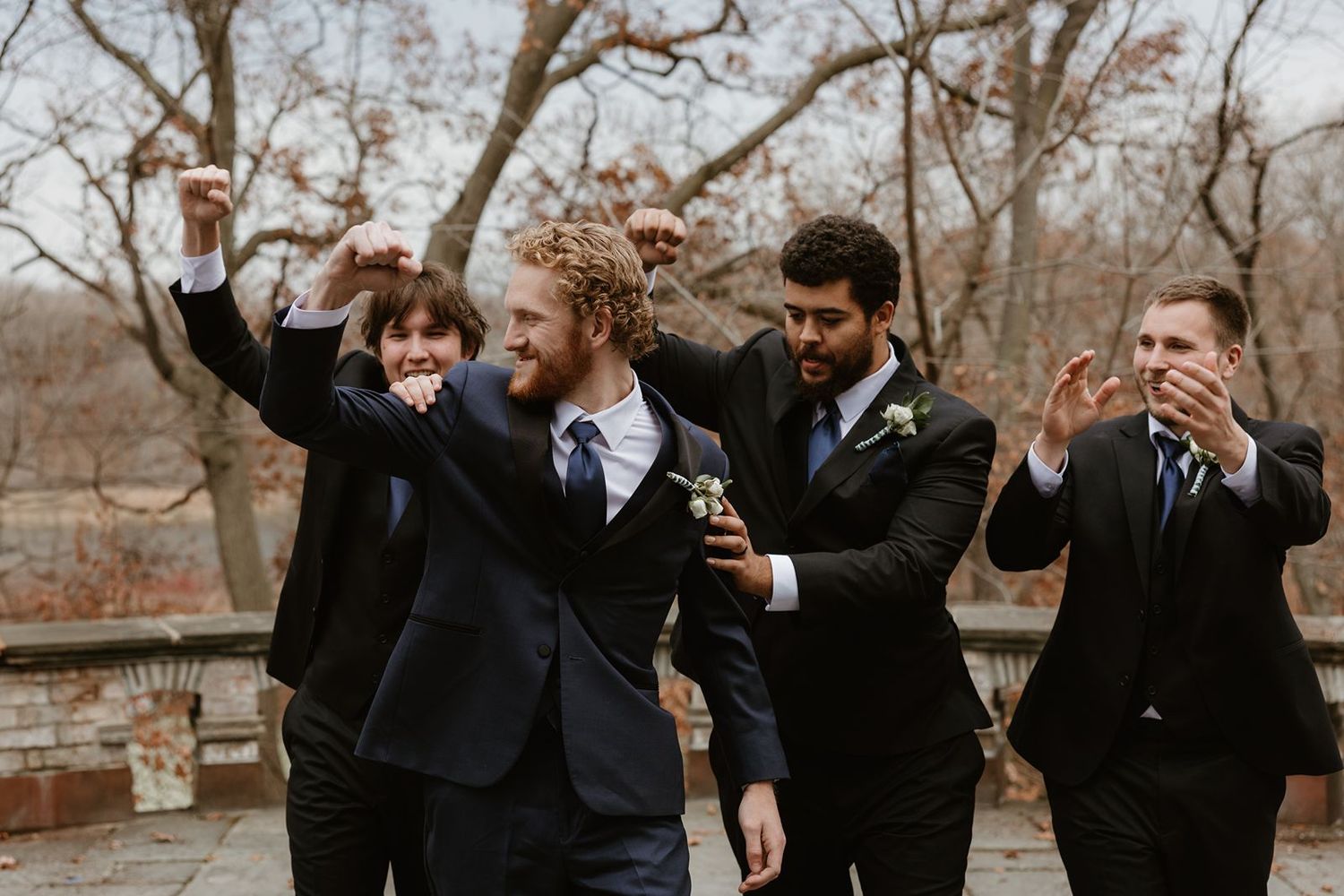 Groomsmen in black suits celebrate joyfully during an autumn wedding.