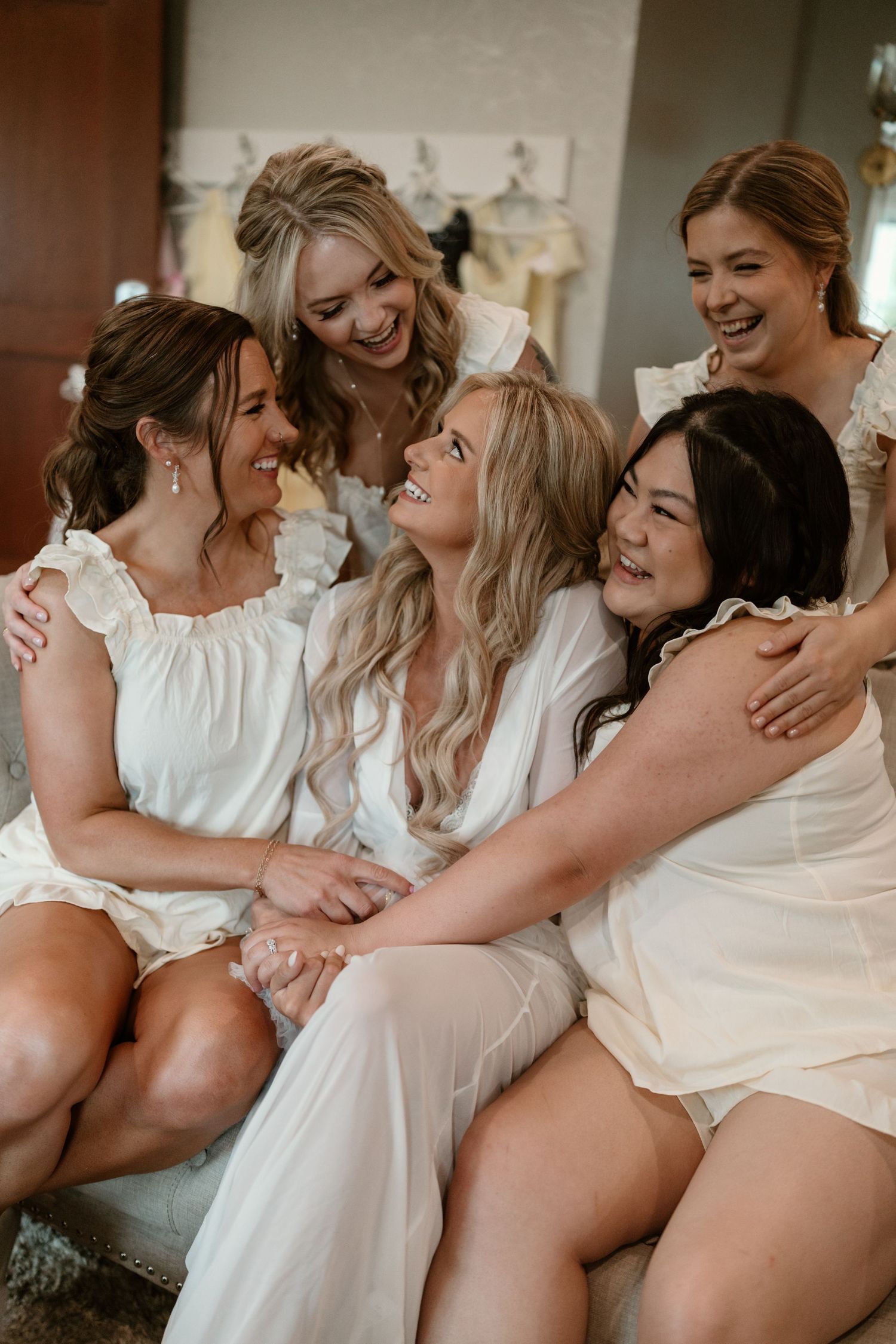A group of women in white dresses laugh and embrace together during a joyful moment at a wedding celebration.