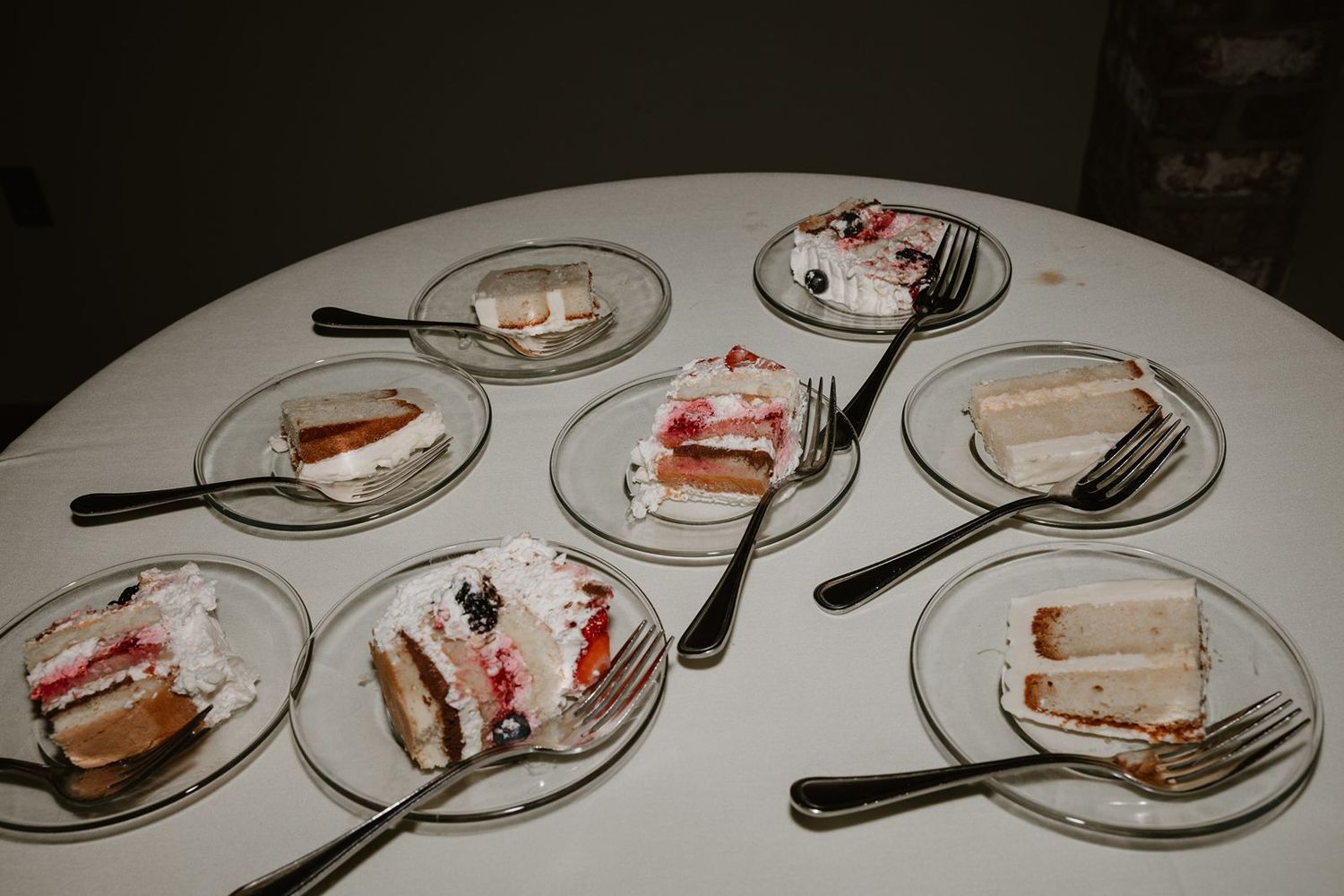 Multiple plates of sliced wedding cake with forks arranged on a round white table.