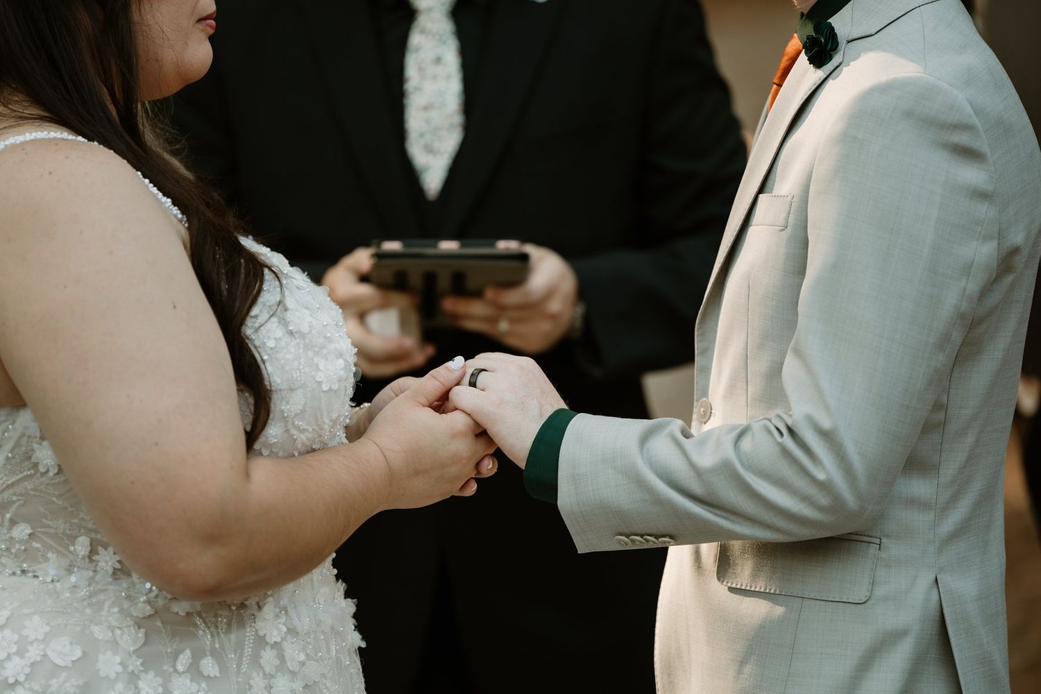 Bride and groom exchanging rings during wedding ceremony, officiant holding book in background.