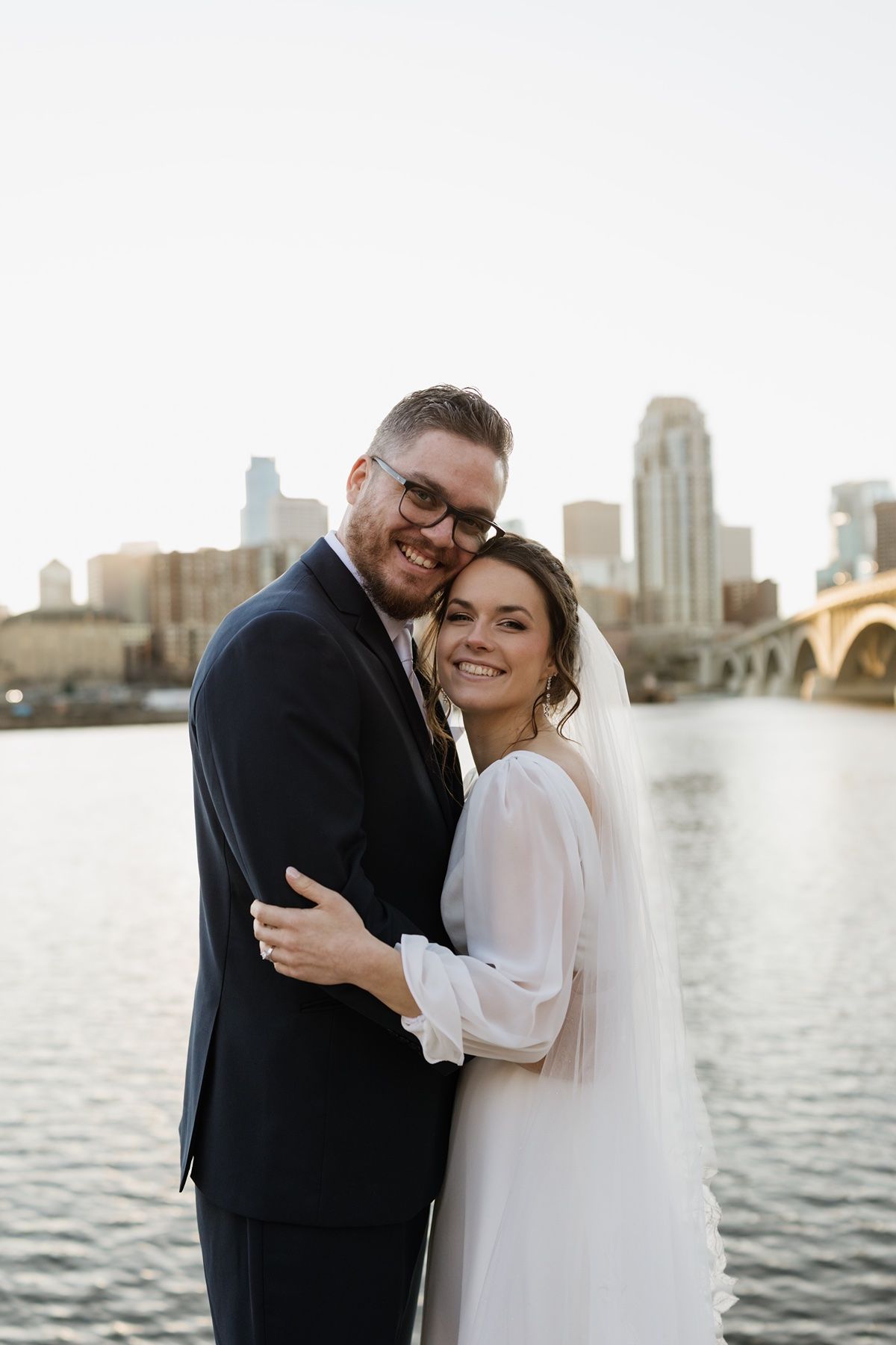 A smiling bride and groom embrace in front of a city skyline and river on their wedding day.