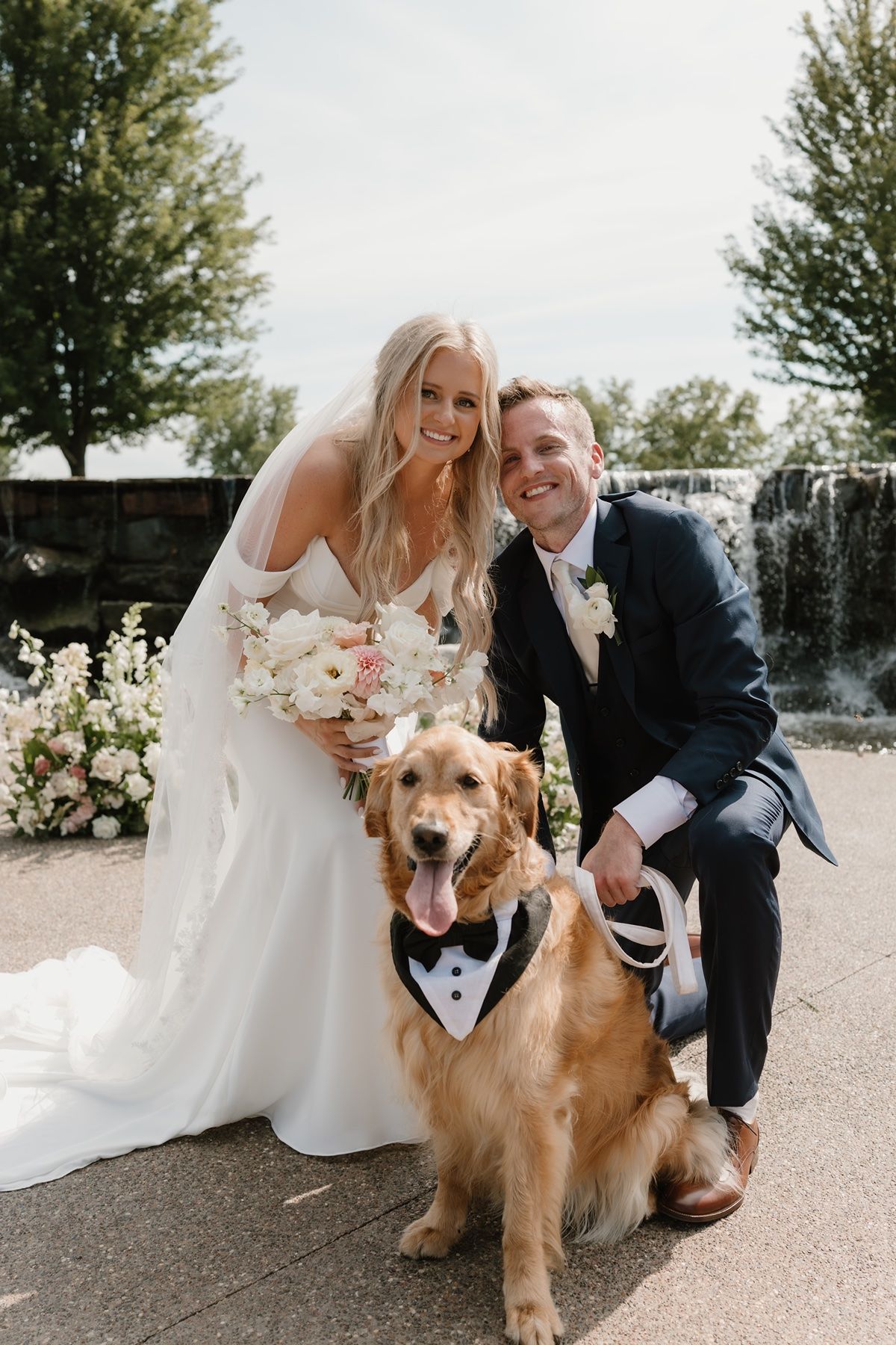 Bride and groom pose with their golden retriever wearing a tuxedo bandana on their wedding day near a fountain.