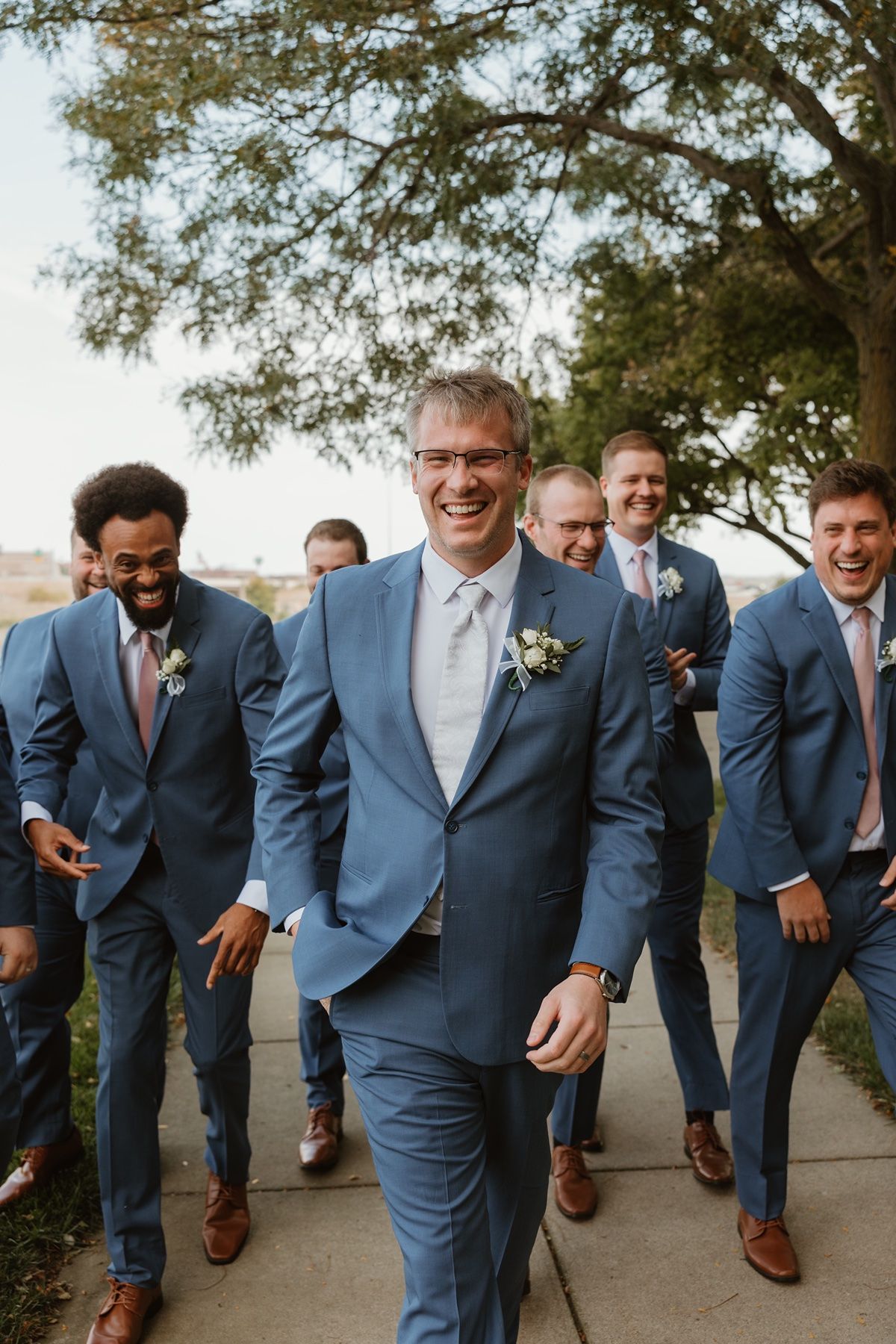 Groom and groomsmen laughing in matching blue suits with pink ties and boutonnieres walking outdoors at a wedding.