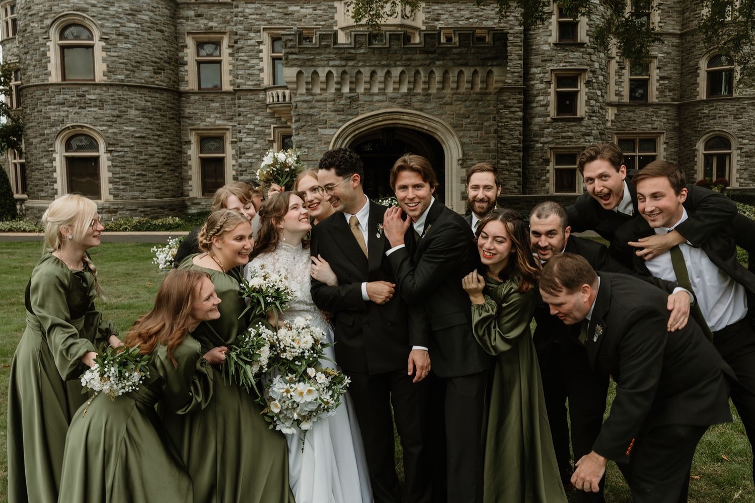 Wedding party in green attire celebrating with bride and groom before a stone castle backdrop.