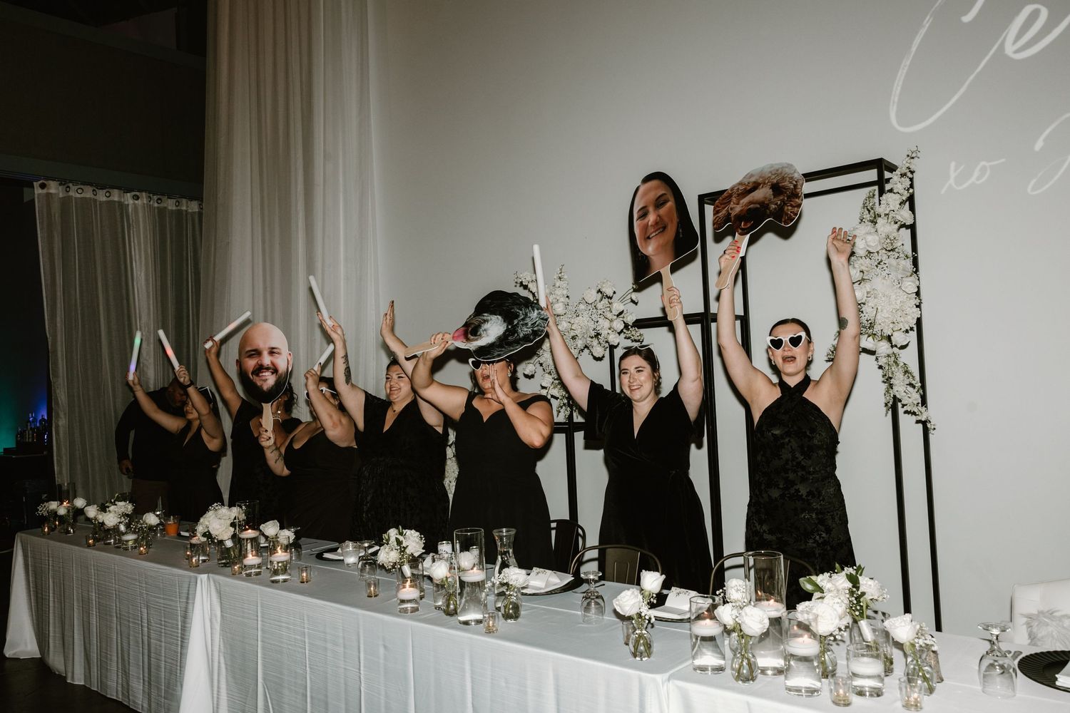 Wedding party dressed in black poses energetically behind a decorated banquet table with candles and floral arrangements.