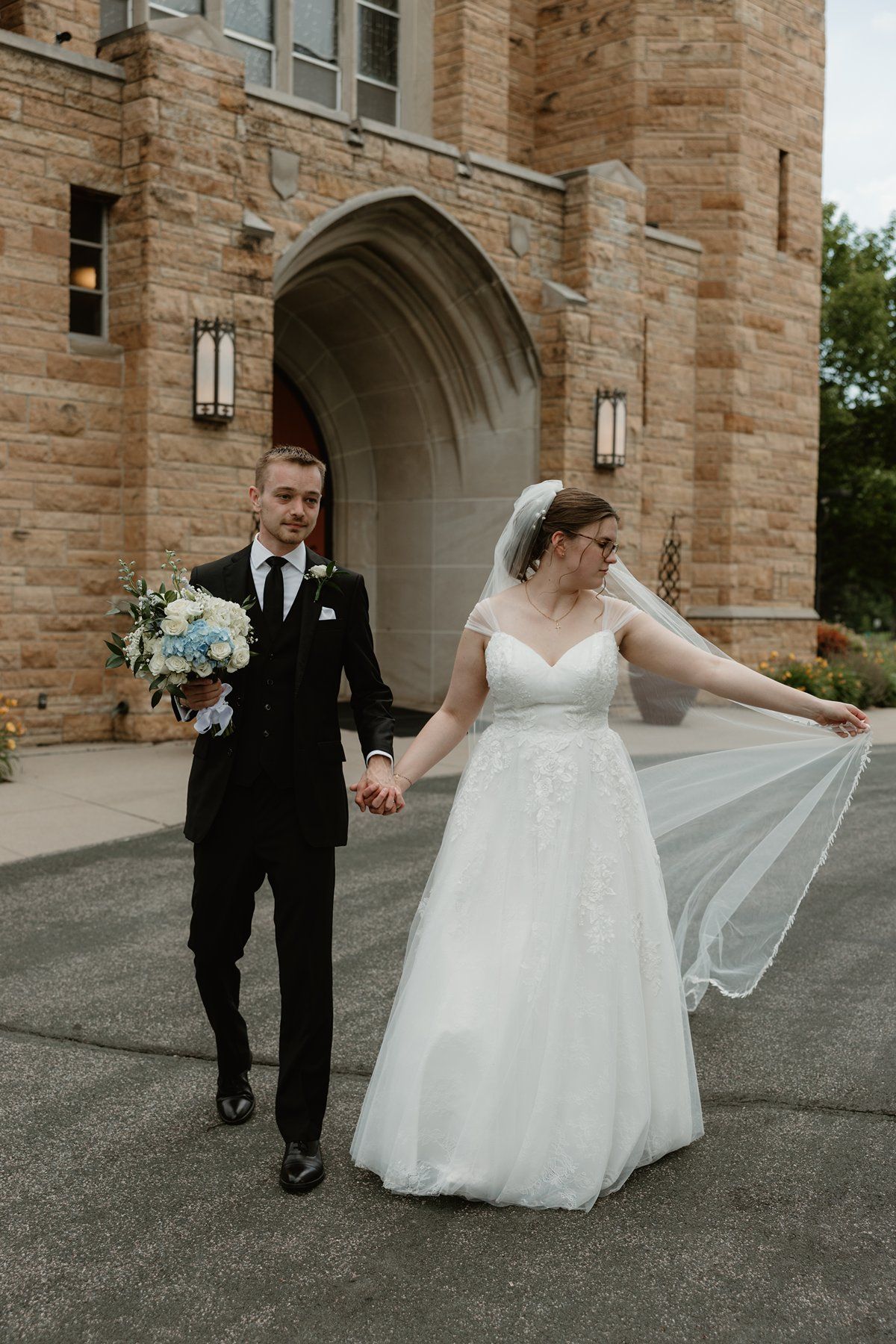 A couple shares a joyful moment as the bride's veil flows gracefully against the stone archway backdrop.