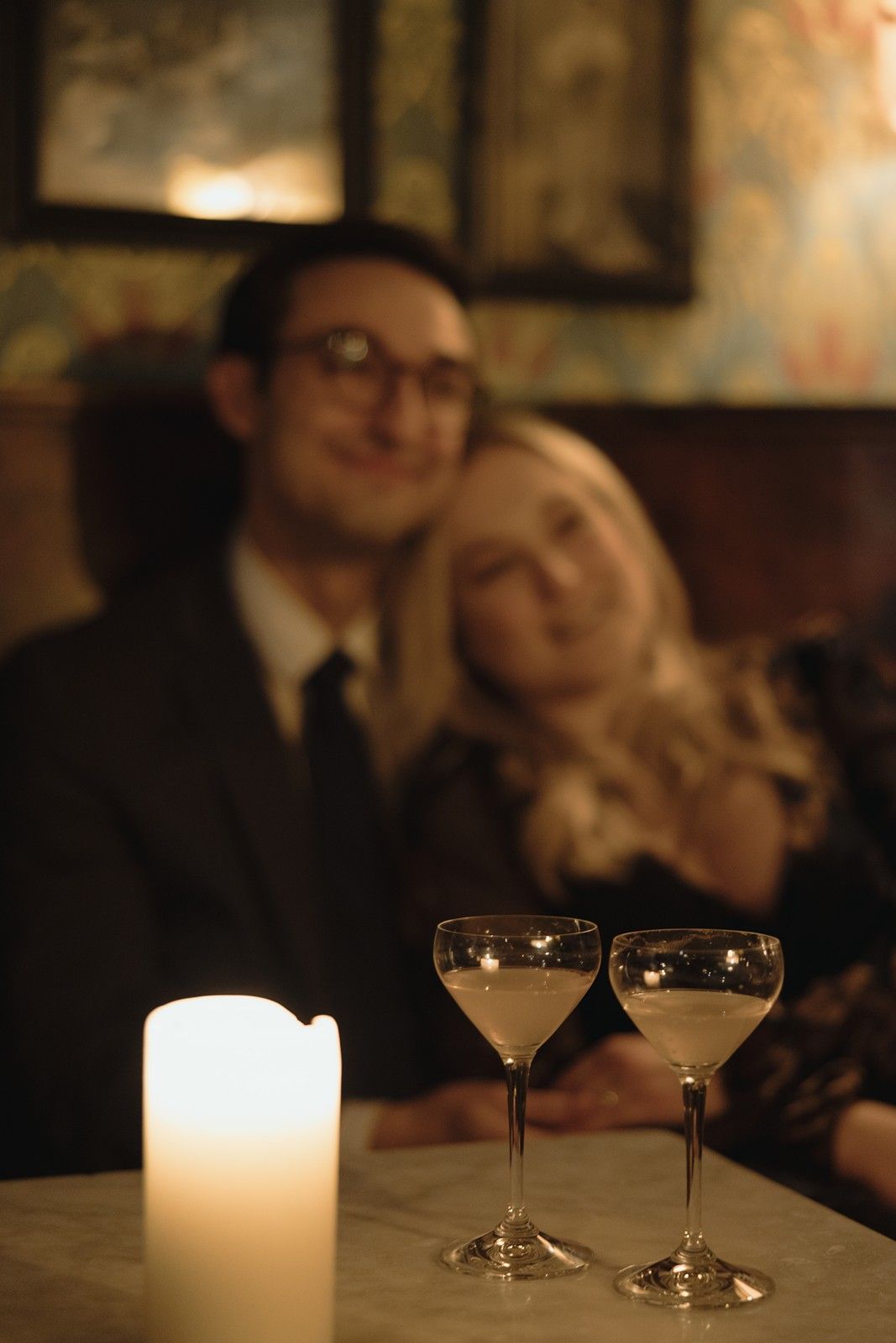 Romantic couple sharing intimate moment at candlelit bar with two martini glasses and glowing candle on table.