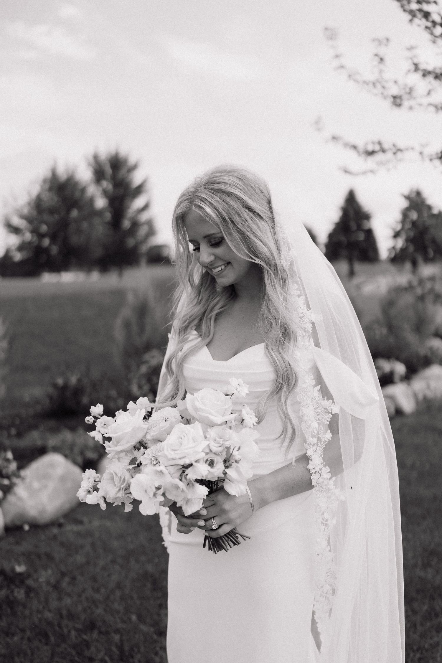 A bride in a flowing white dress and veil holds a delicate bouquet of white flowers in an outdoor garden setting.