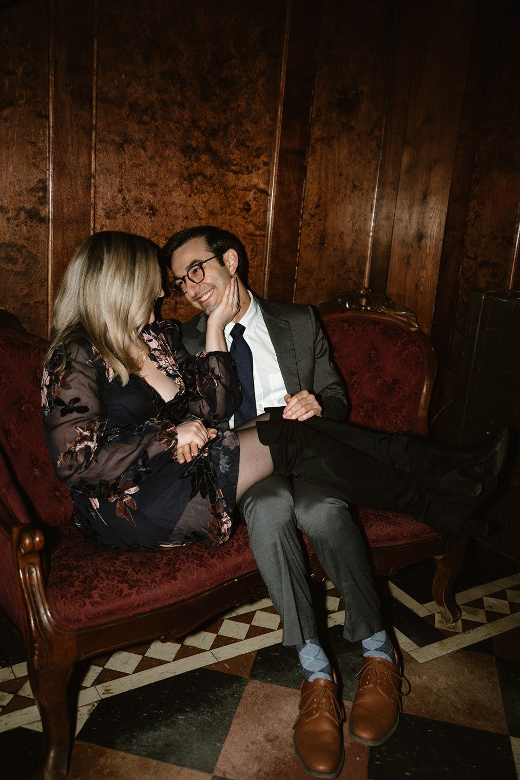 A couple sits closely together on a vintage red velvet couch against dark wooden paneling in moody lighting.