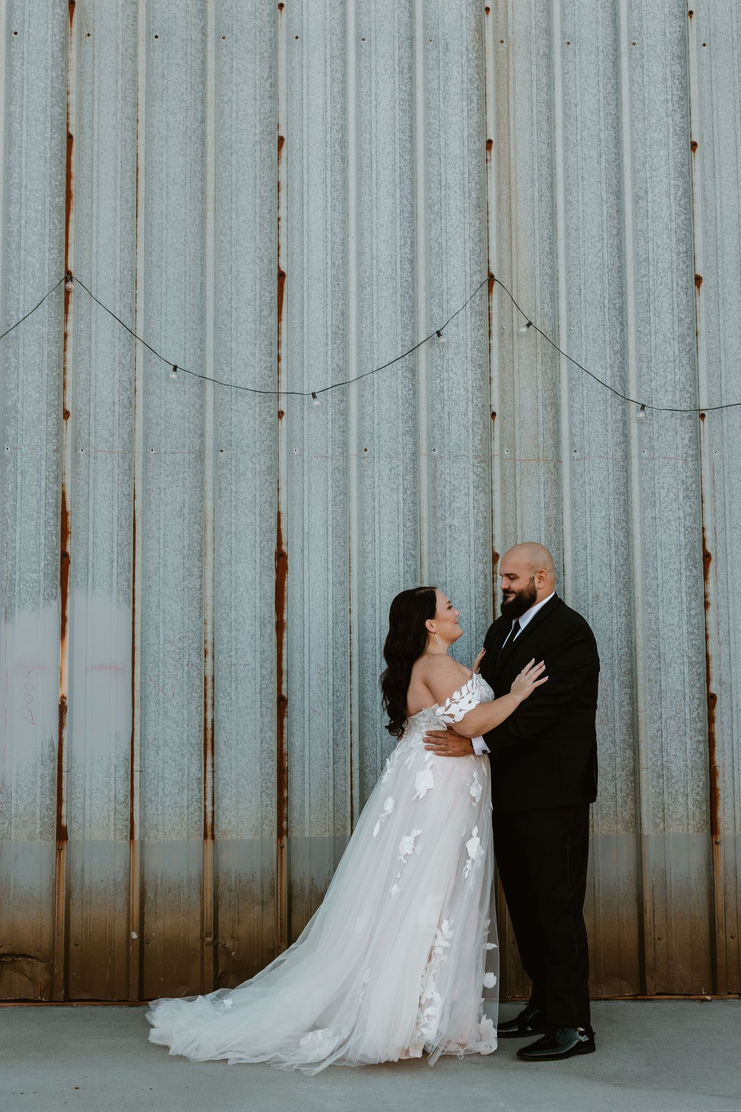 Wedding couple in elegant attire embrace against industrial corrugated metal wall backdrop creating moody ambiance.