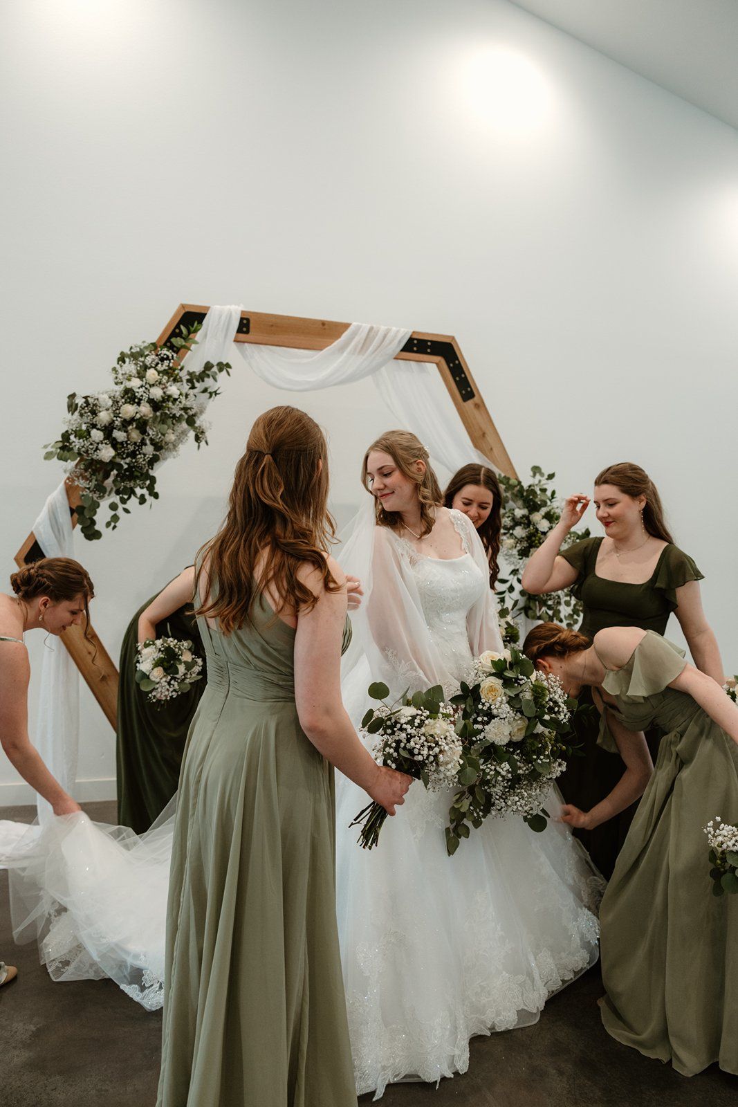 A wedding party in sage green bridesmaid dresses gather around the bride in a white gown with floral decorations.