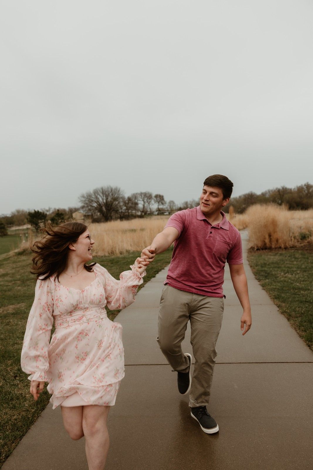 A couple holding hands while joyfully walking together on a concrete path through a grassy outdoor area.