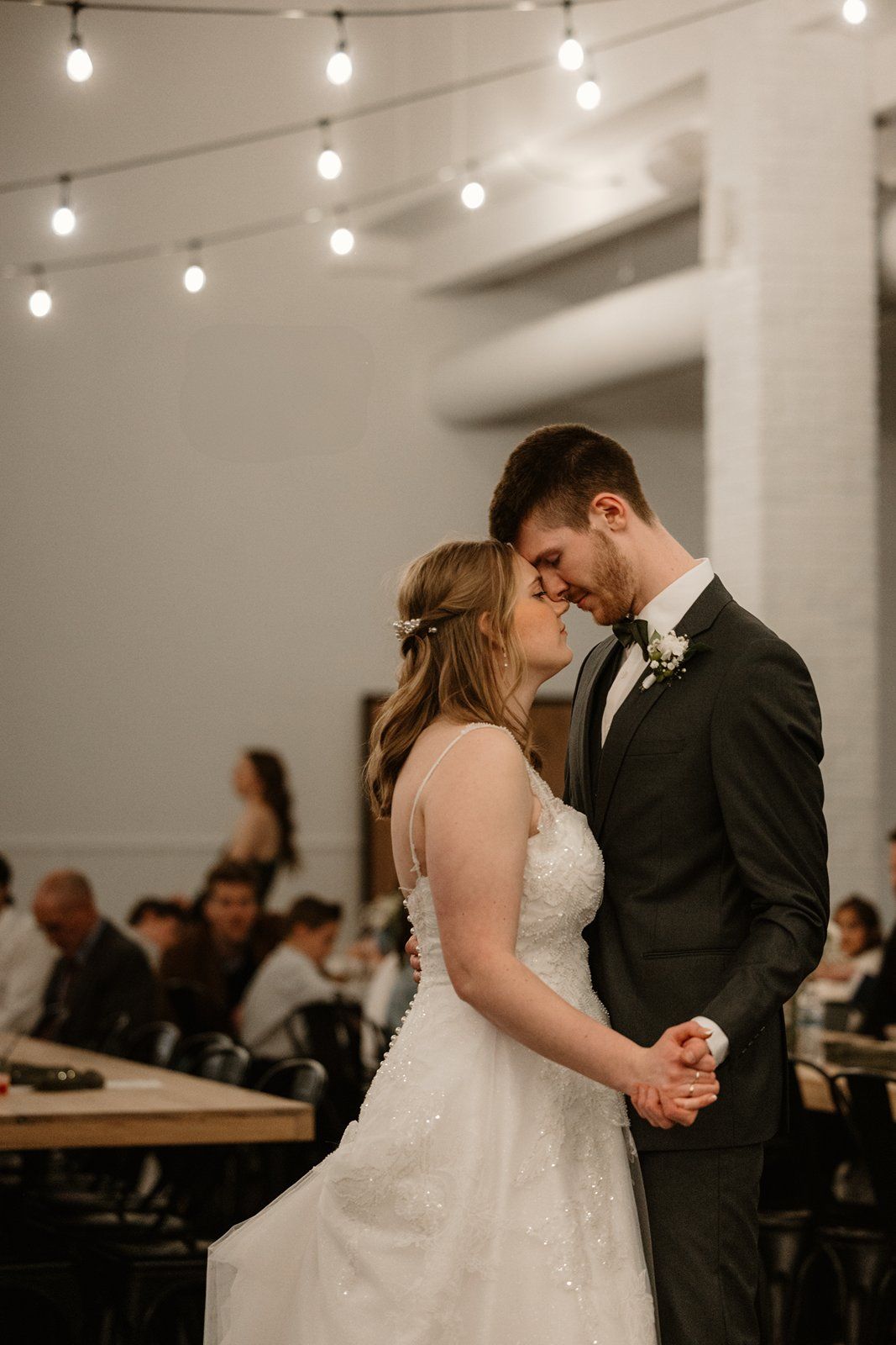 Couple sharing an intimate dance moment under string lights at an indoor wedding venue.