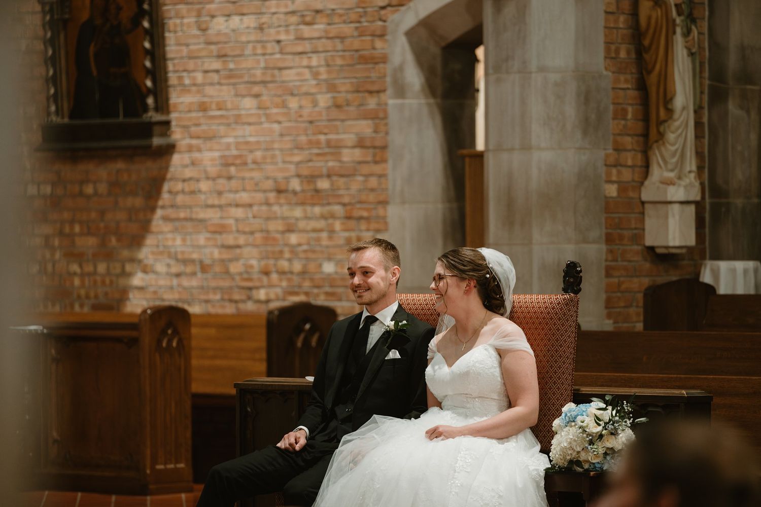 Couple seated on wooden pews inside historic brick church with wedding attire and floral bouquet.