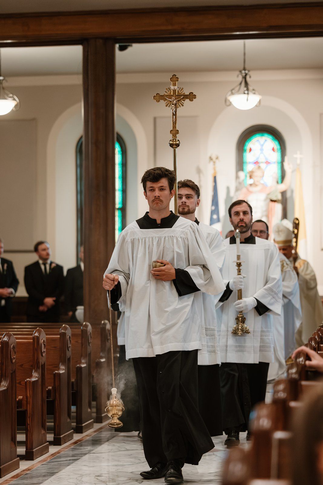 Altar servers in white robes walk down the aisle of a traditional Catholic church during a ceremony.