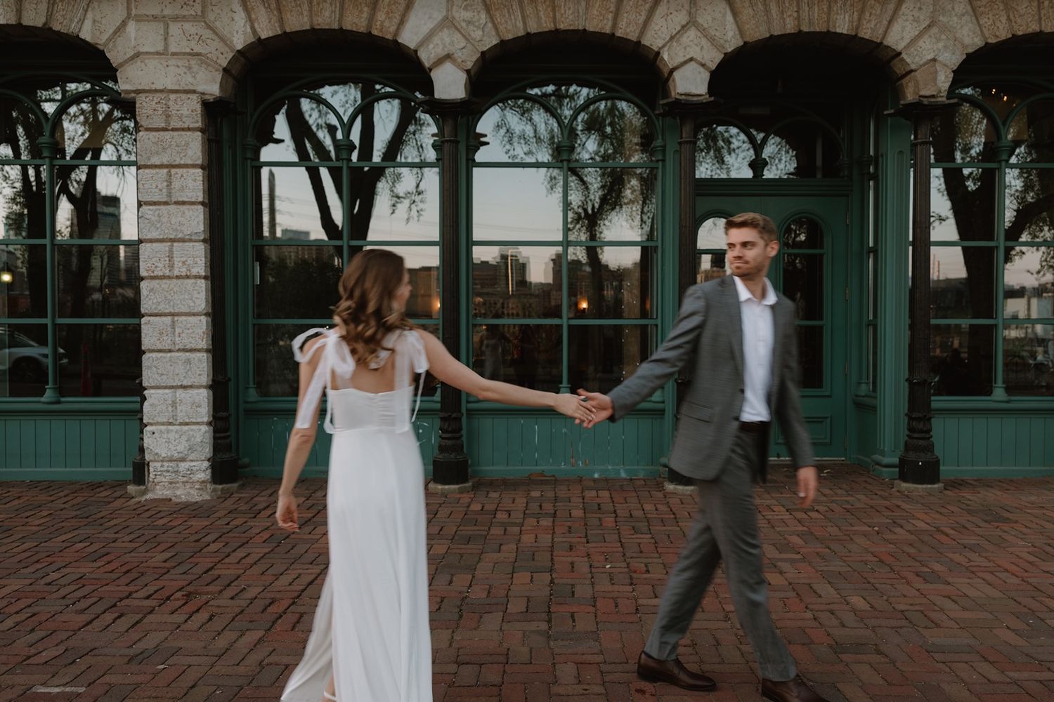 A bride and groom hold hands walking on brick pavement outside a building with green arched windows.