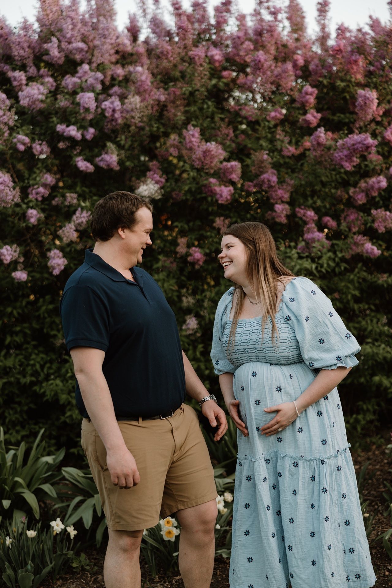 Smiling pregnant couple standing in front of blooming pink flowers, woman in blue floral dress cradling baby bump.